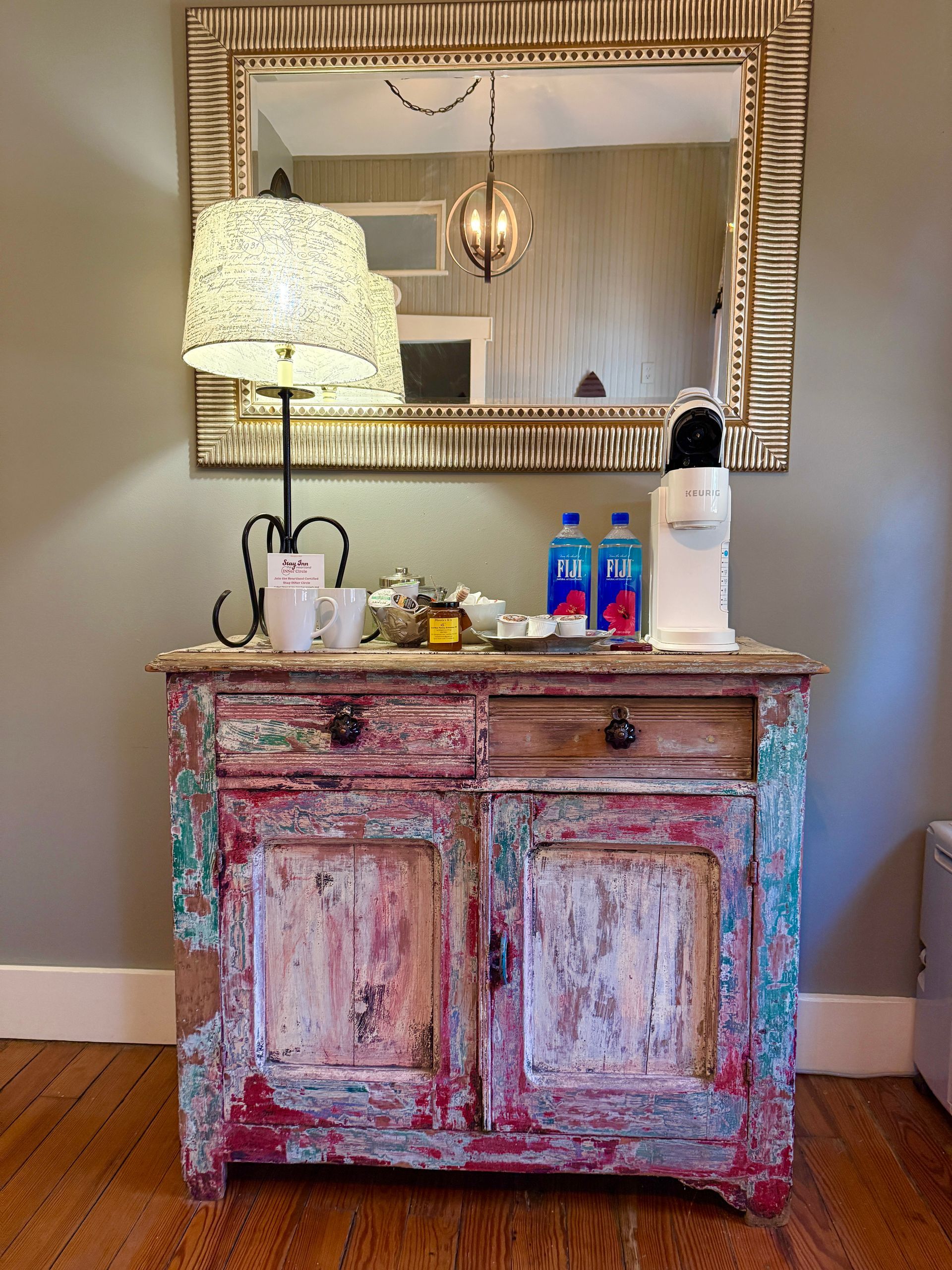 A decorative cabinet with coffee supplies, a lamp, and a mirror with a decorative frame.