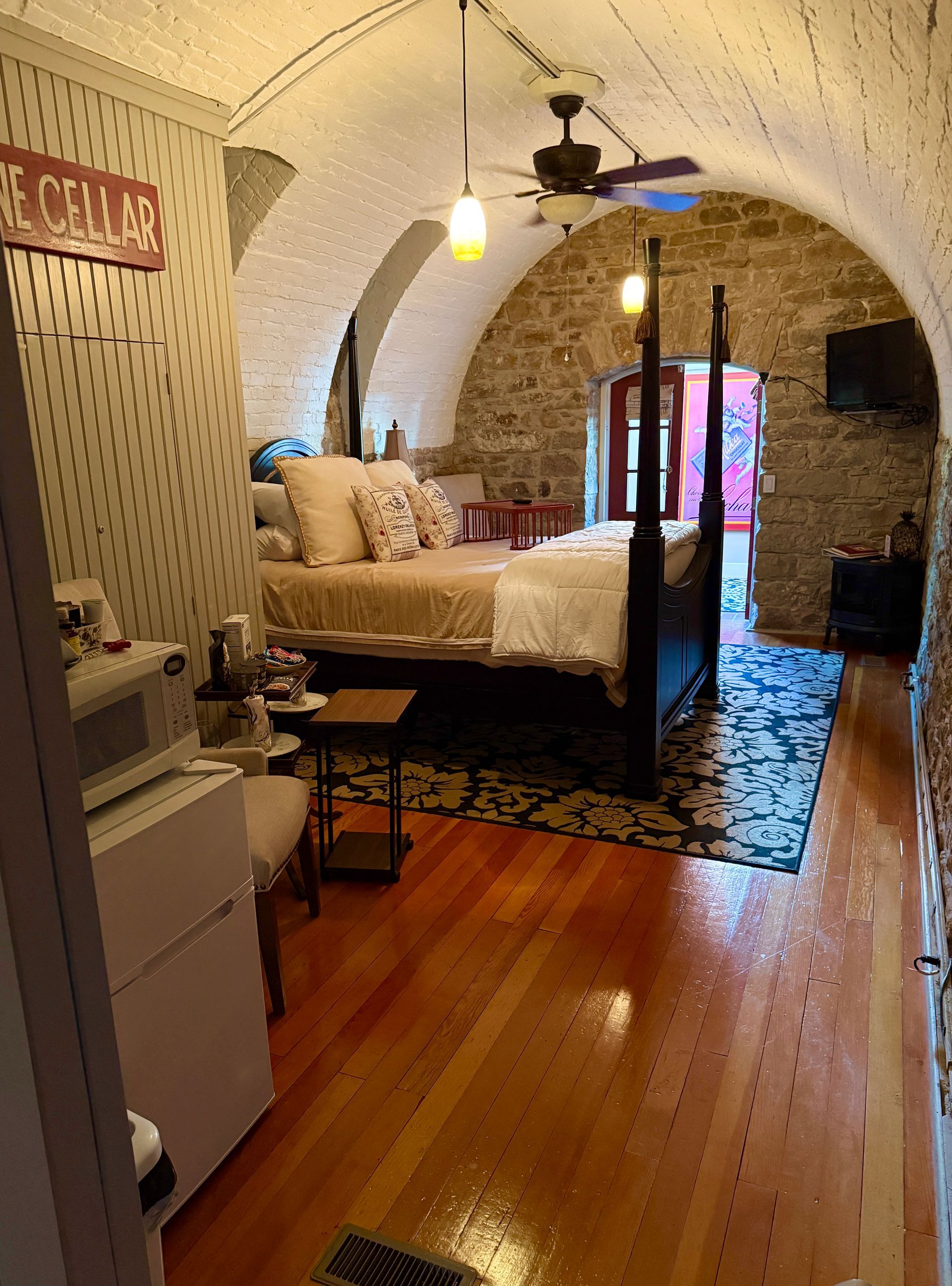 Bedroom with arched stone walls, wood floors, four-poster bed, rug, ceiling fan.