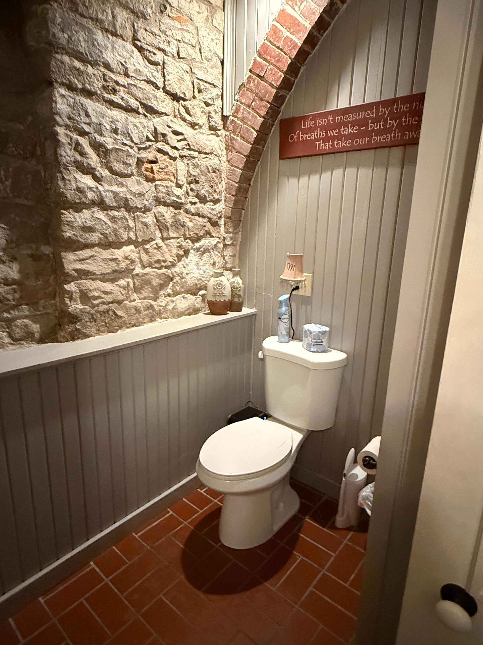 Small bathroom with stone wall and brick arch, toilet, wainscoting, and brick floor.