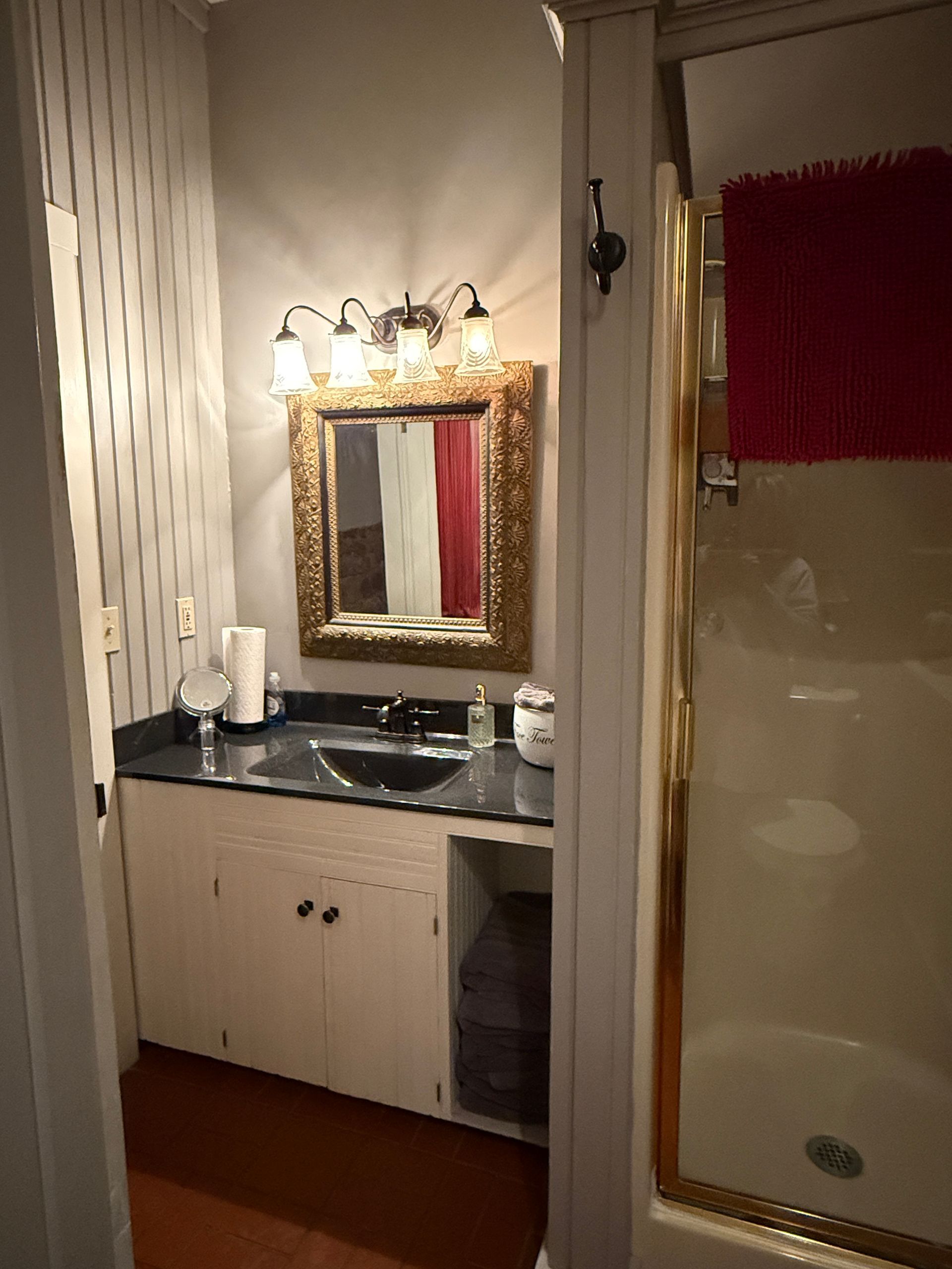 Small bathroom with a vanity, mirror, and shower. Gold accents, red rug, white and black.