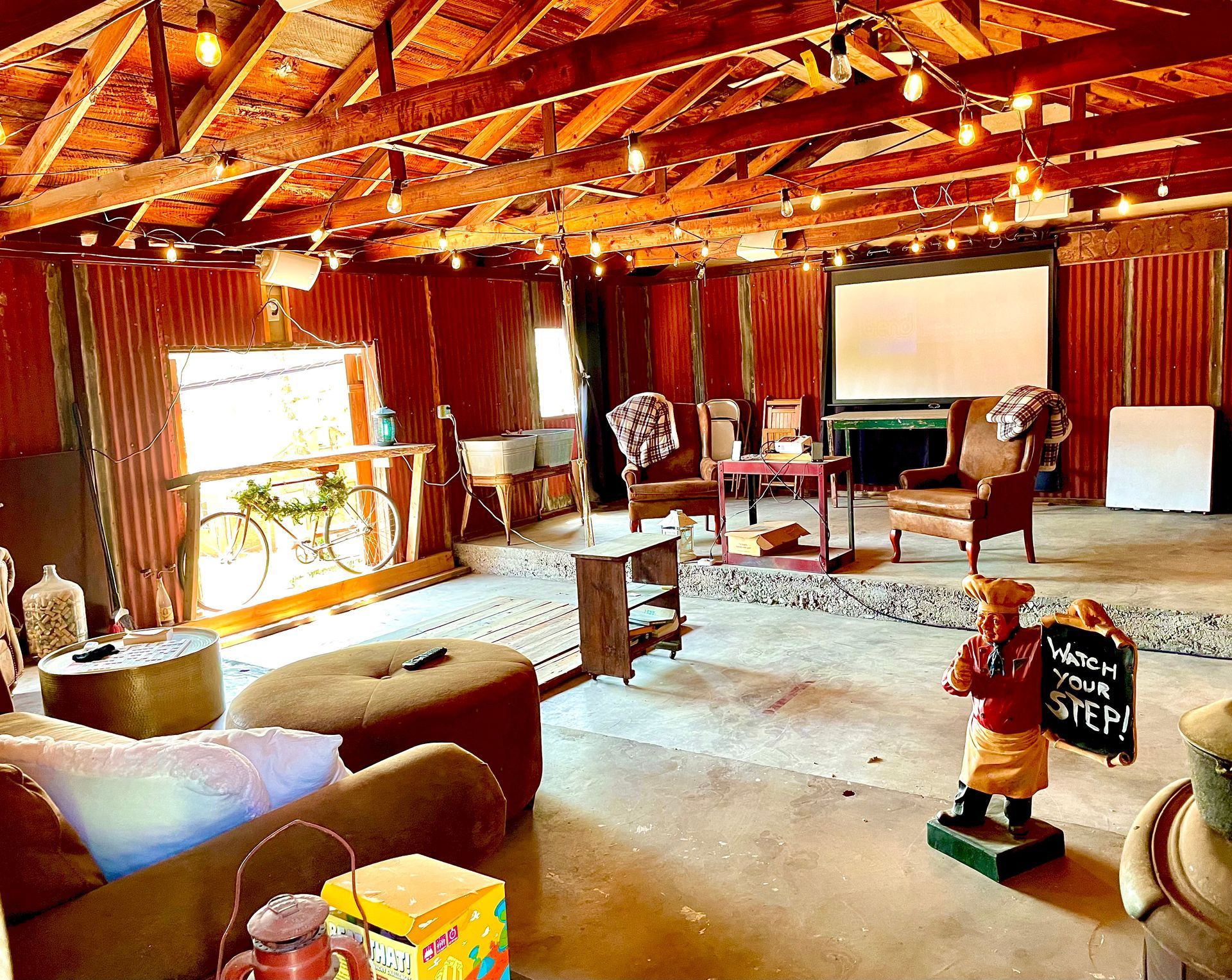 Rustic interior with furniture, string lights, and a projector screen. A sign holder figurine stands in front.