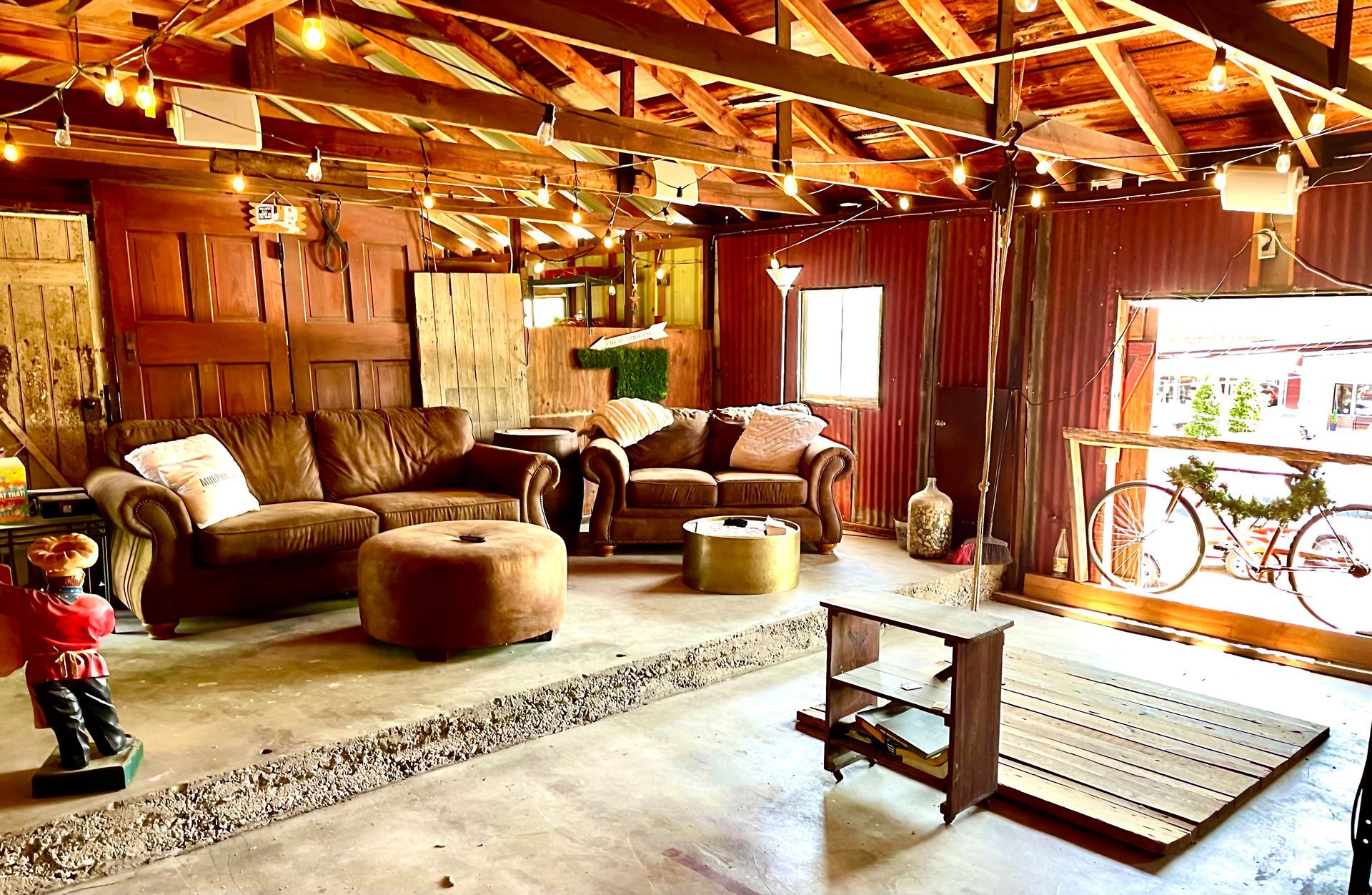 Cozy indoor space with couches, ottoman, and string lights; barn-like structure with concrete floor and doorway.