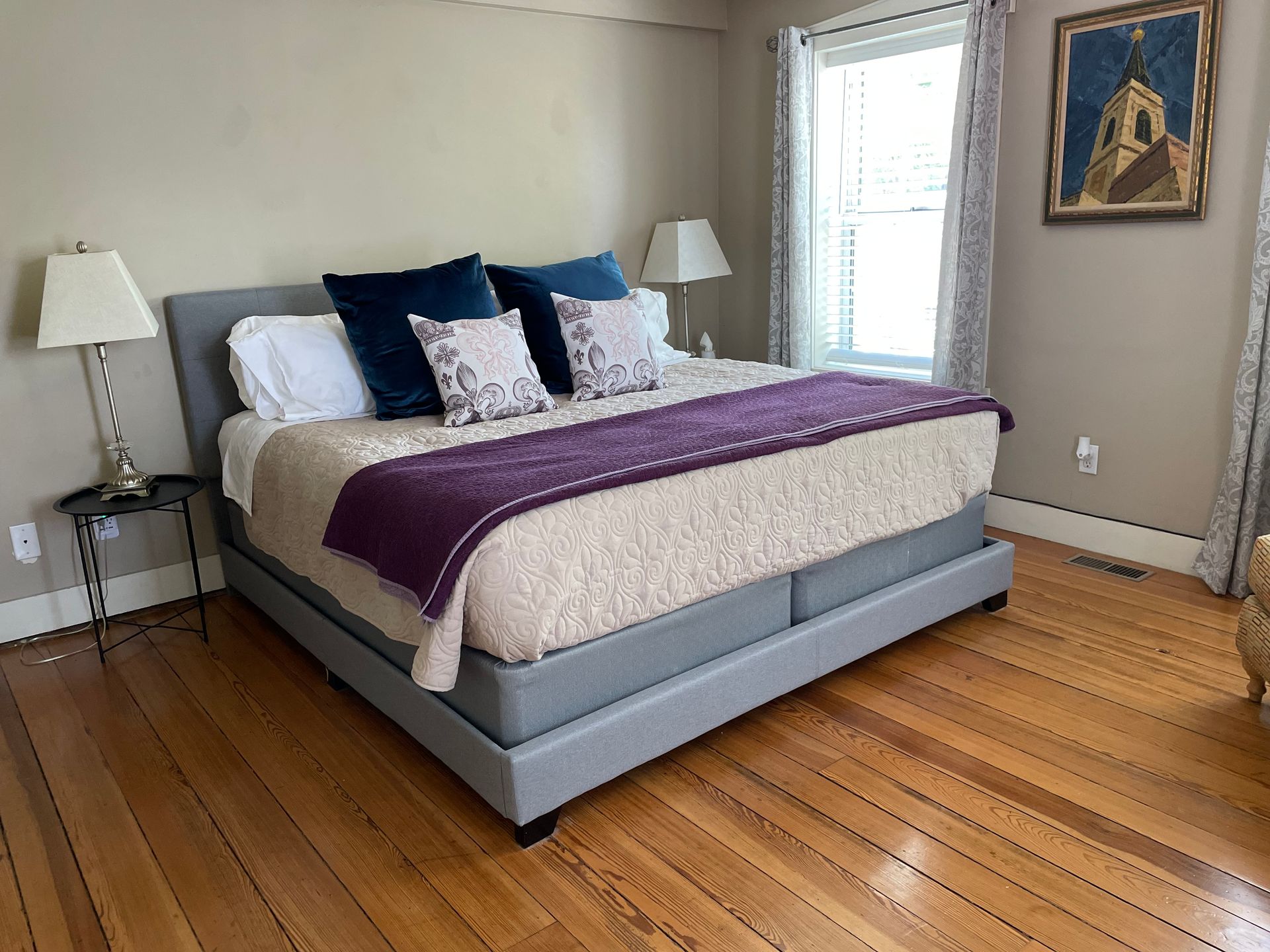 Bedroom with king-sized bed, grey upholstered frame, white bedding, purple throw, blue and patterned pillows, wood floor.