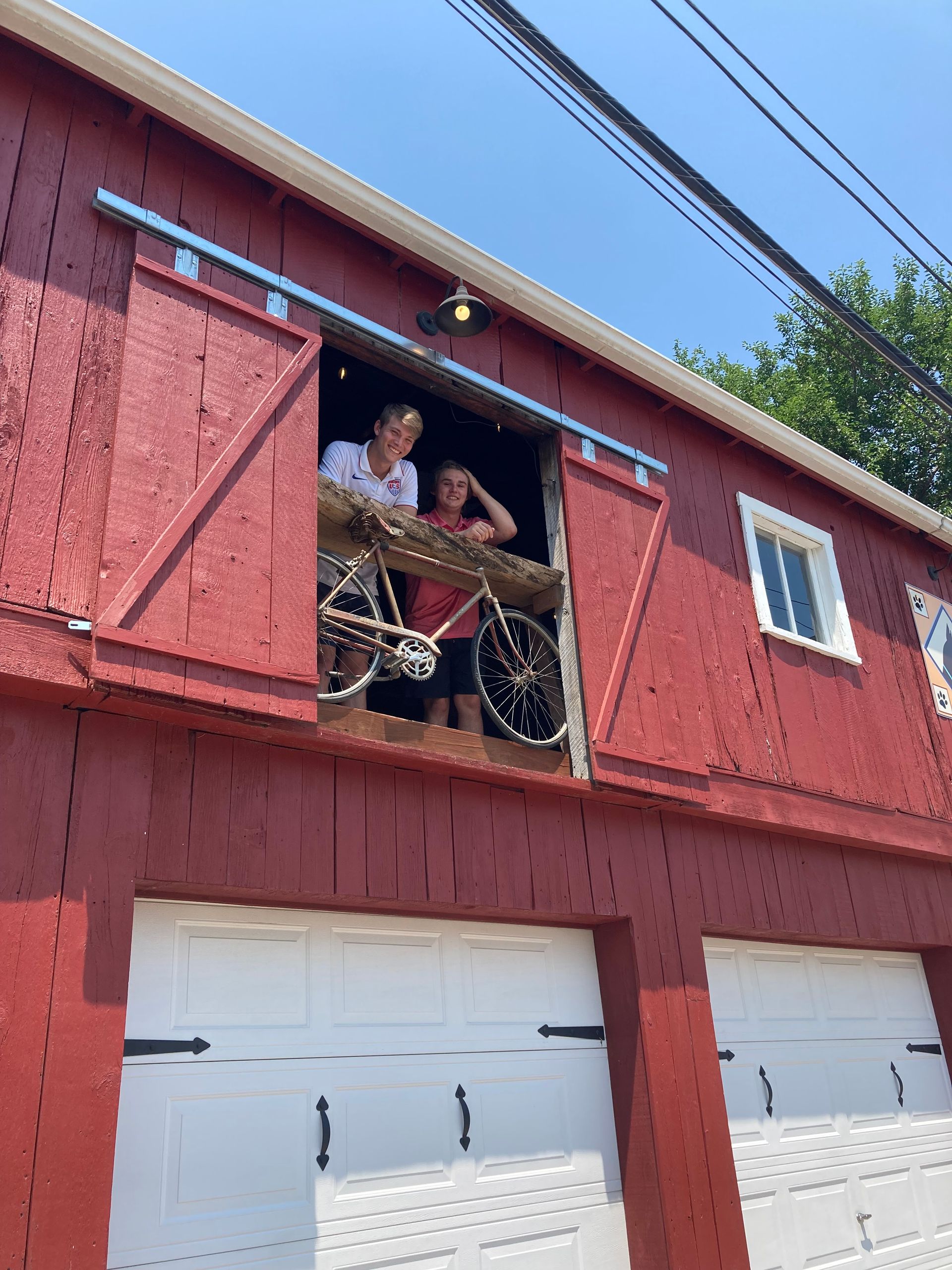 Red barn with garage doors below, two people looking out a window with a bicycle.