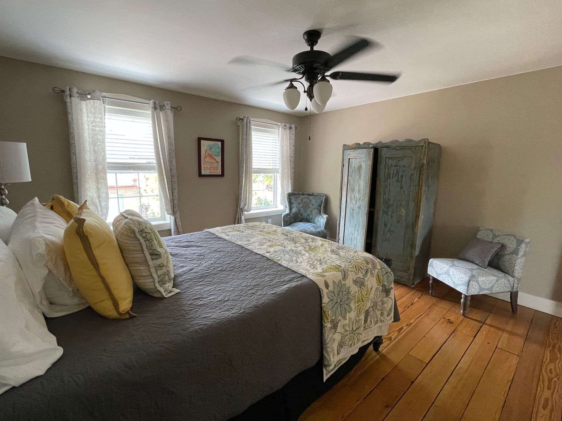 Bedroom with bed, armoire, chairs, windows, and ceiling fan. Wooden floor, neutral walls, and natural light.