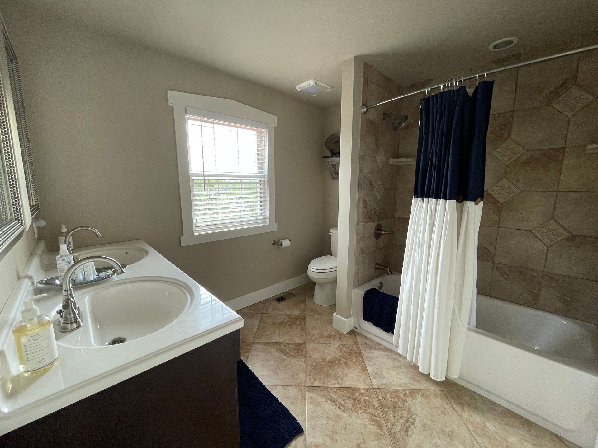 Bathroom with tan-tiled floor, double sink, window, toilet, and a bathtub with a blue and white shower curtain.