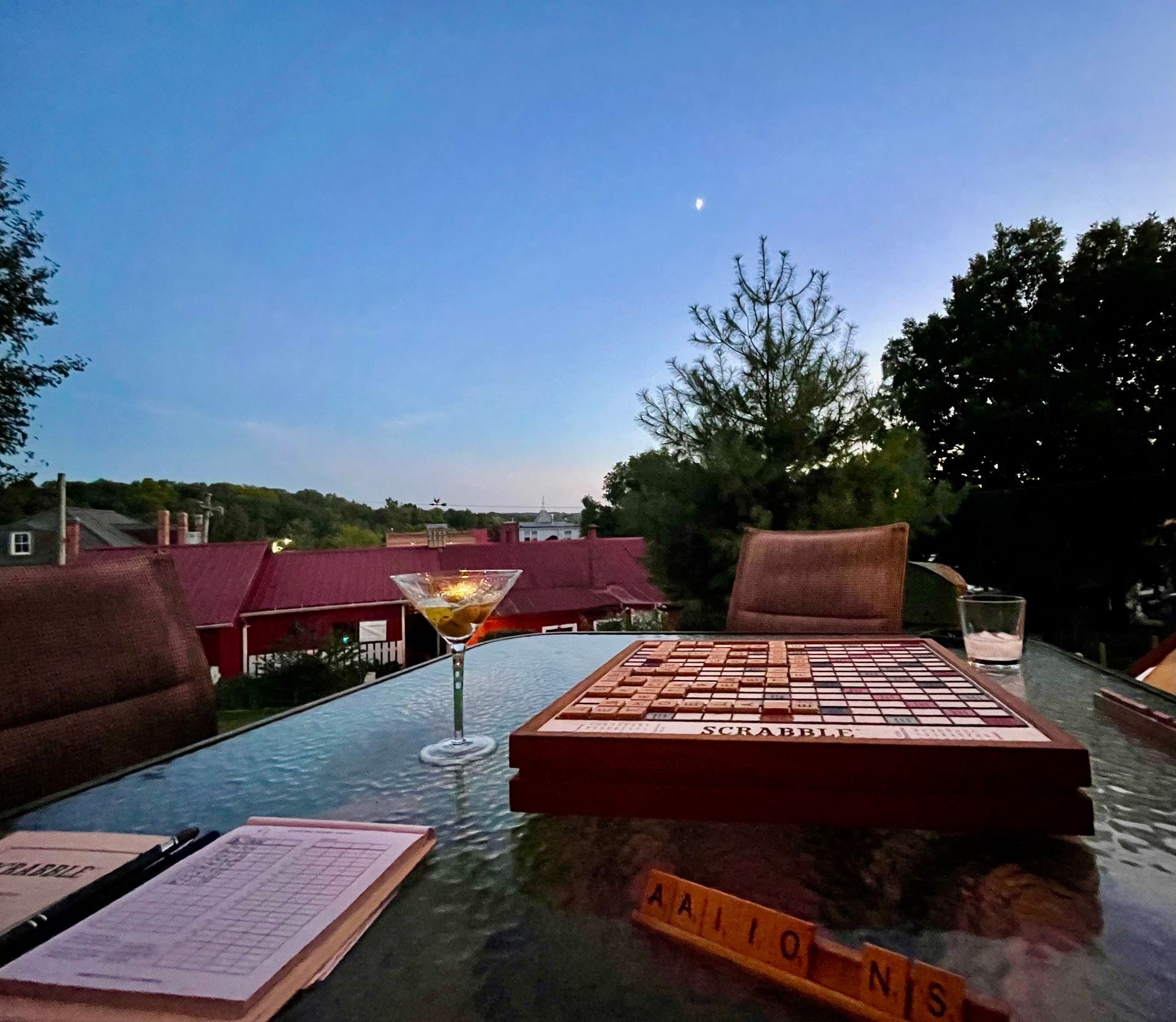 Scrabble game on a glass table with cocktail, overlooking a town at dusk.