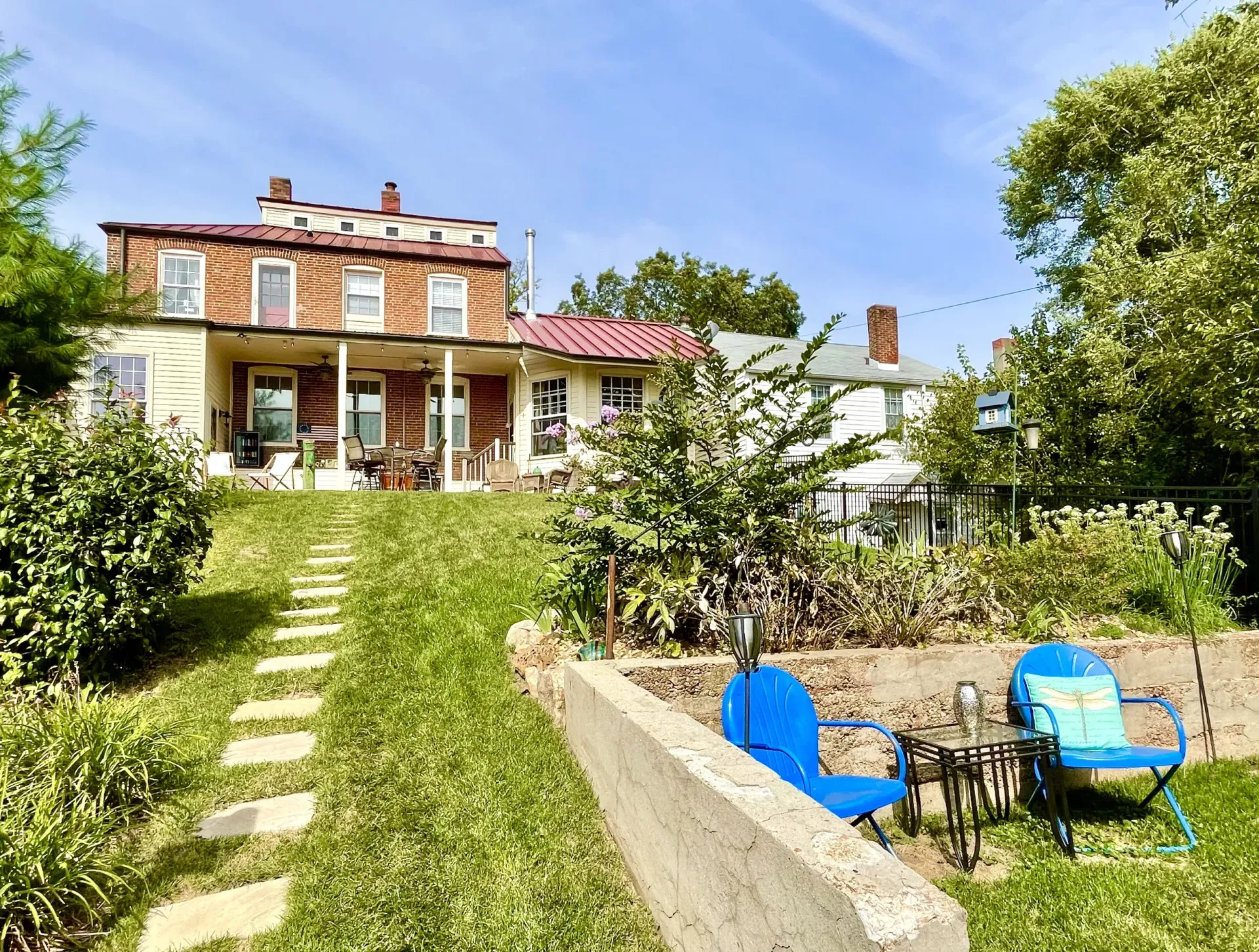 A two-story brick house with a red roof sits on a slight hill, with a stone path and chairs in a garden.