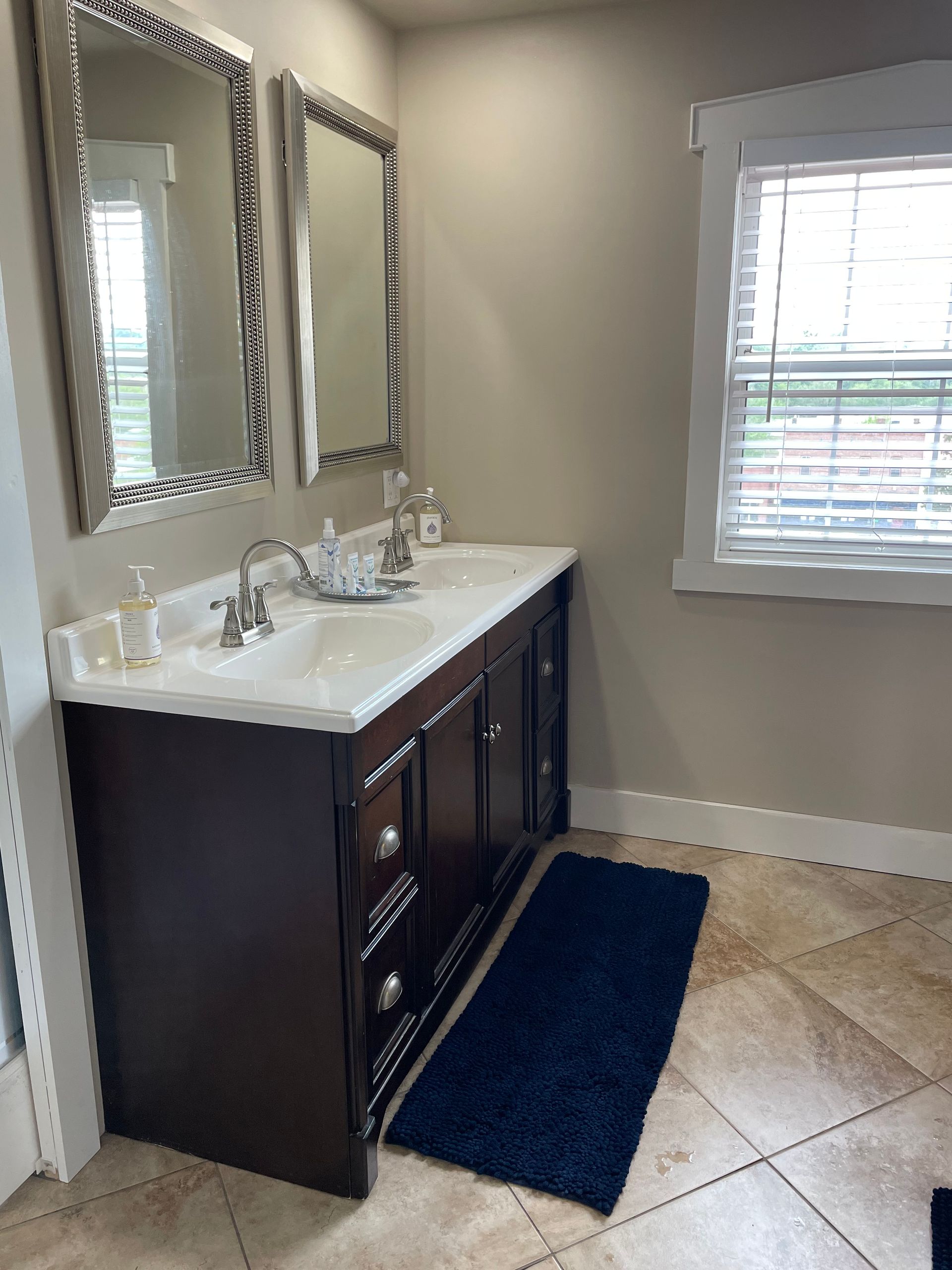Bathroom with dark brown vanity, two mirrors, and a blue rug on tiled floor.