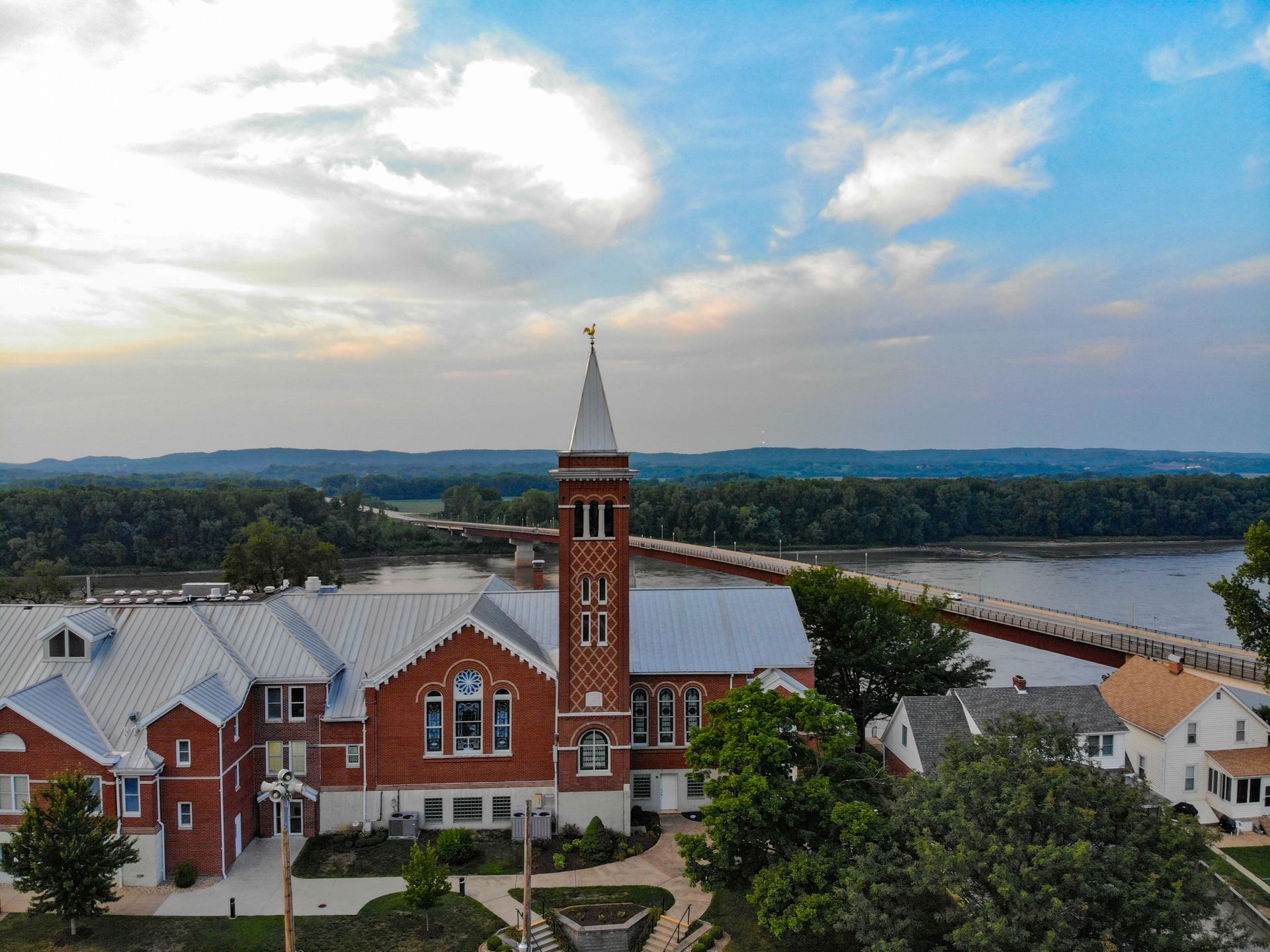 Red brick church with tall steeple, river, and bridge under cloudy sky.