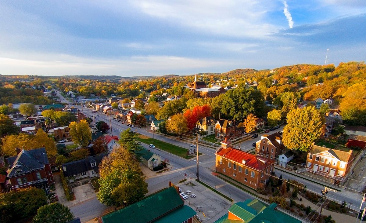 Aerial view of a town with autumn foliage. Buildings line streets, trees show red and gold leaves under a blue sky.