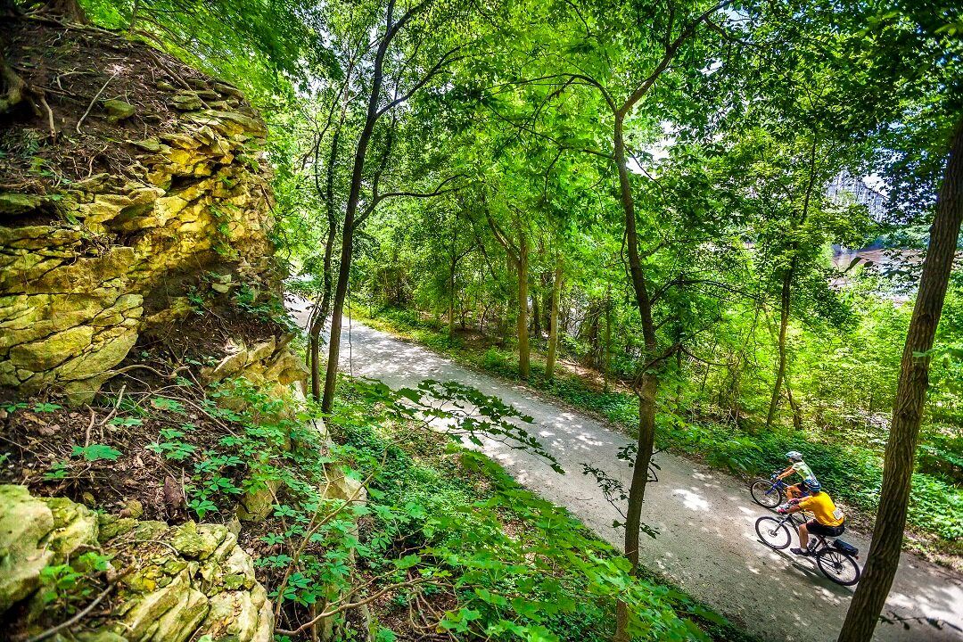 Cyclist riding on a gravel path through a lush forest, next to a rock cliff face.