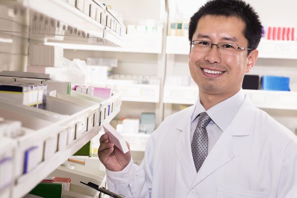 A smiling pharmacist is standing in front of a shelf in a pharmacy.
