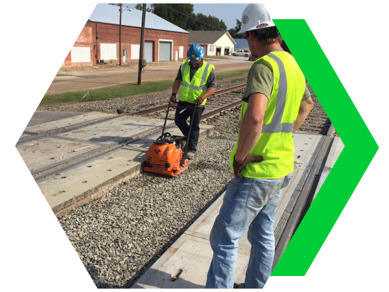 Two workers in yellow vests using a machine on a gravel railway crossing near houses