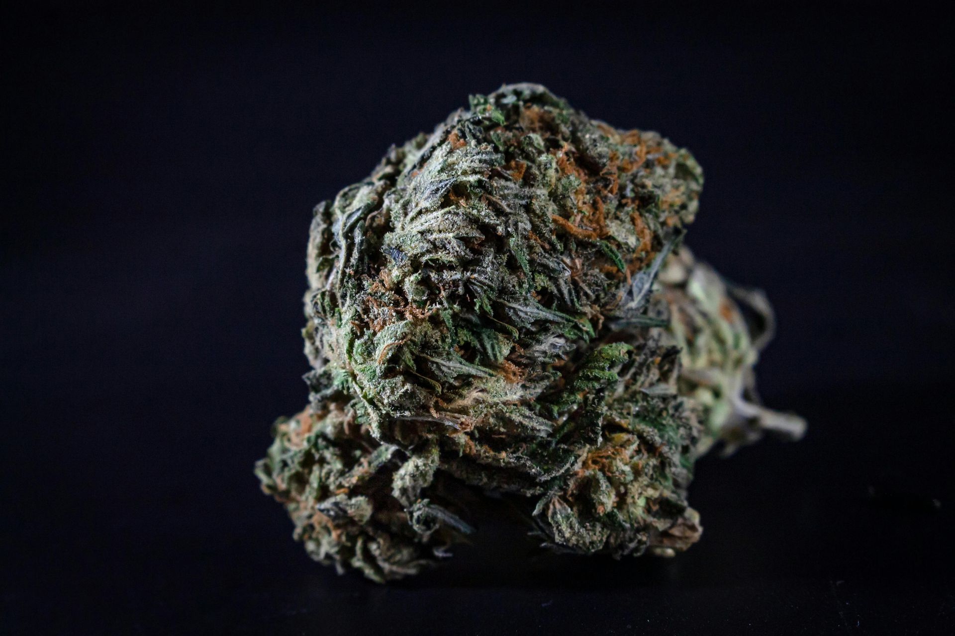 A close-up of a dried cannabis flower bud with green and brown leaves against a black background.