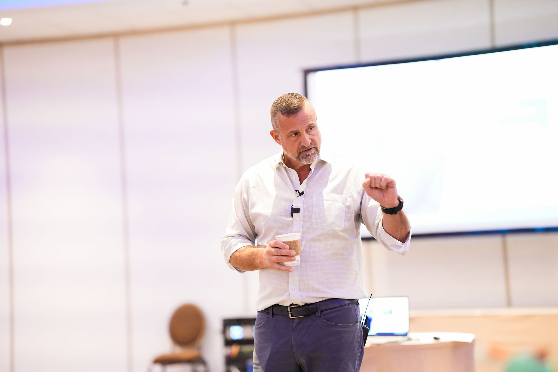 A man is giving a presentation in a conference room while holding a cup of coffee.