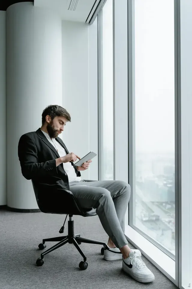 A man is sitting in an office chair looking out of a window while using a tablet.