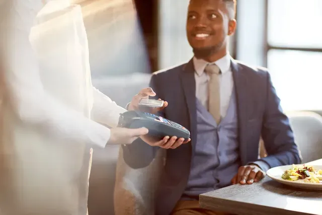 A man in a suit is sitting at a table in a restaurant paying with a credit card.