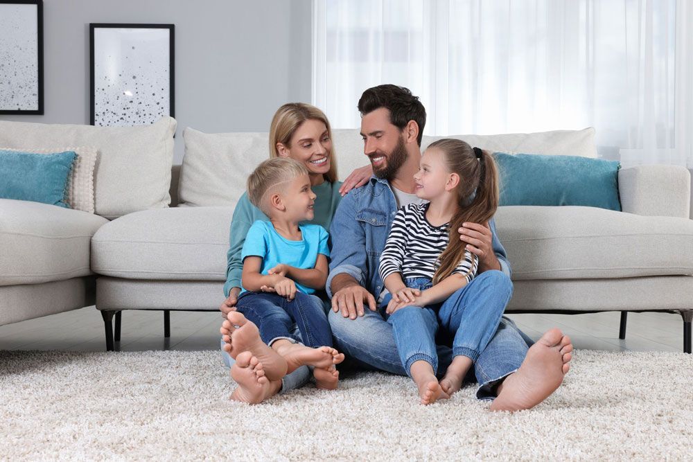 Family of four sitting on a rug in front of a couch, smiling at each other.