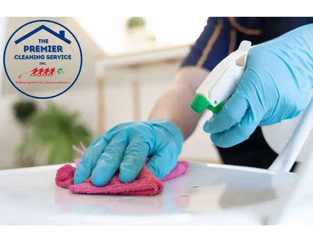 A gloved hand wipes a white surface with a pink cloth, using a spray bottle. Cleaning service logo visible.