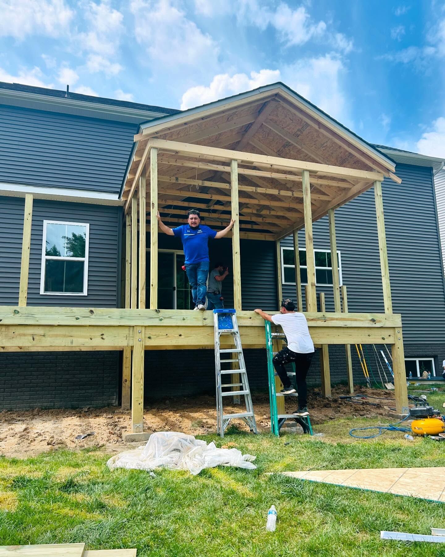 Construction workers building a wooden porch roof on a house, one stands on the deck, two on ladders, sunny day.