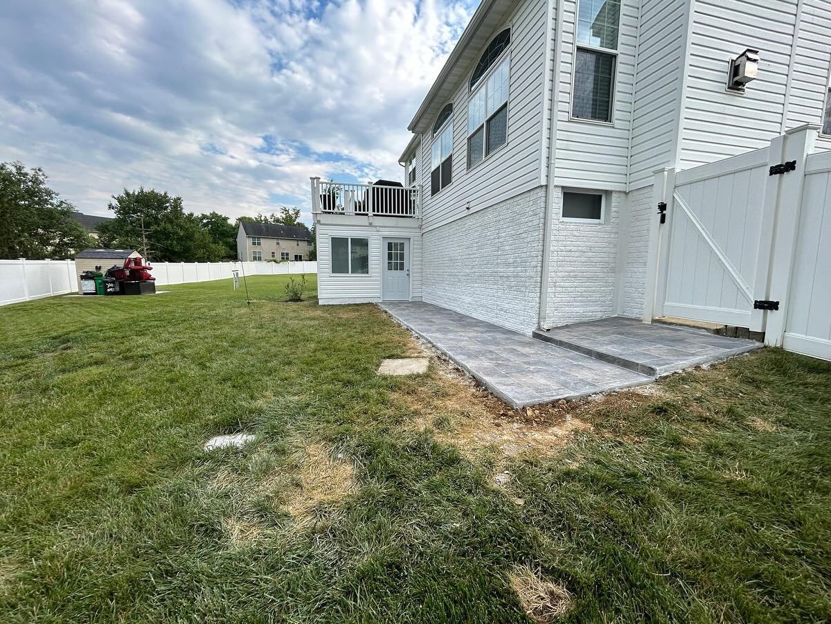 Backyard with white house, patio, and grass. White fence, deck, and overcast sky.