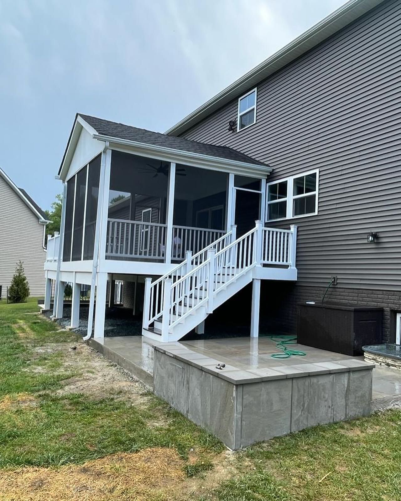 Screened-in porch attached to a house with white railings, stairs, and a concrete patio on a cloudy day.