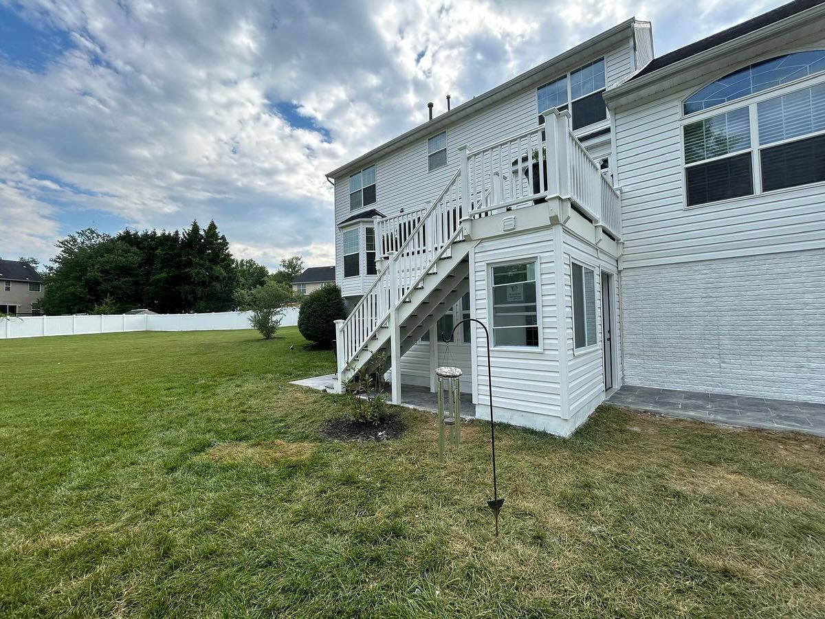White two-story house with a wooden deck and stairs leading to a backyard with green grass and a cloudy sky.