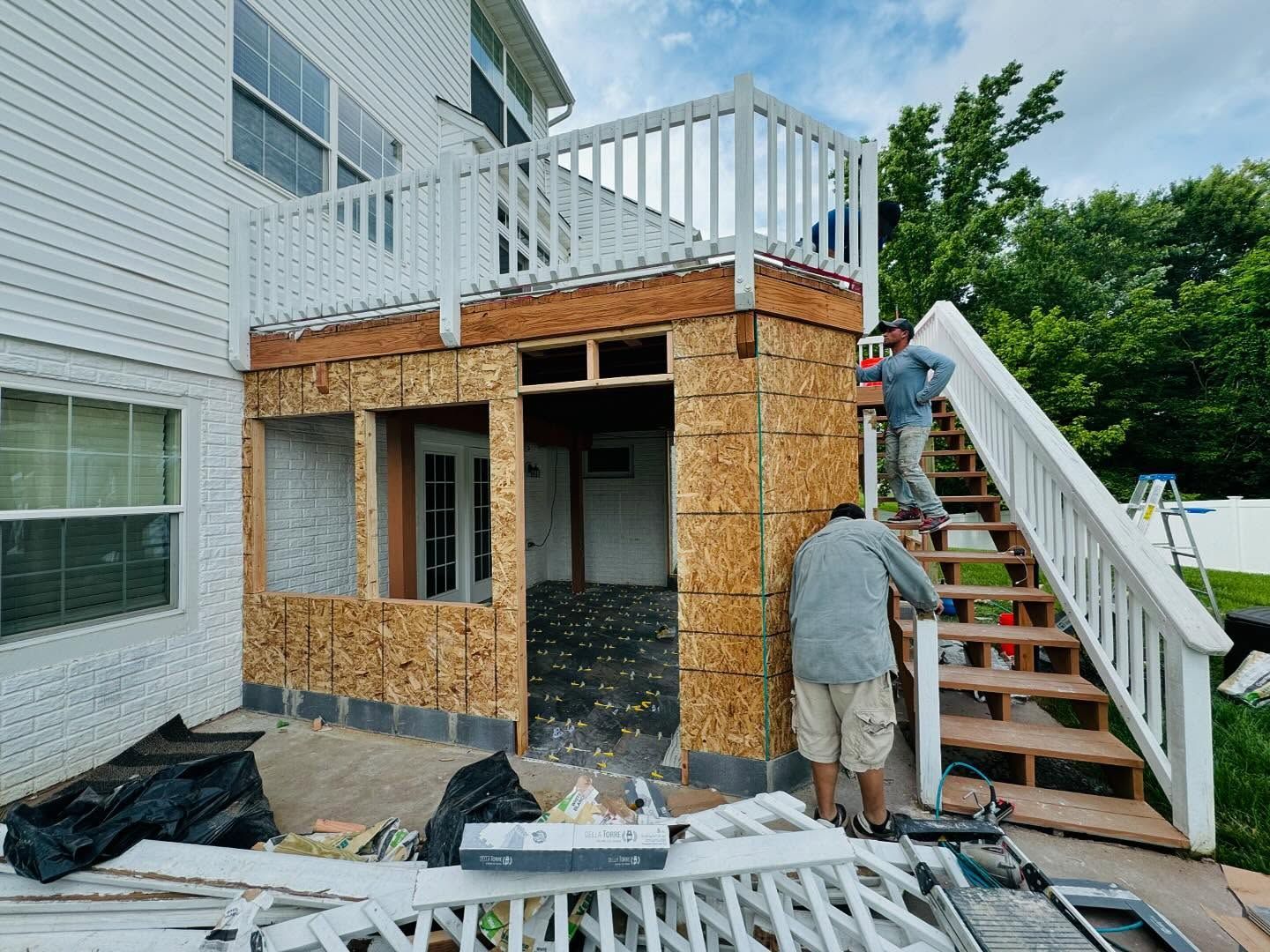 Two men building an outdoor structure attached to a house with a deck and stairs.