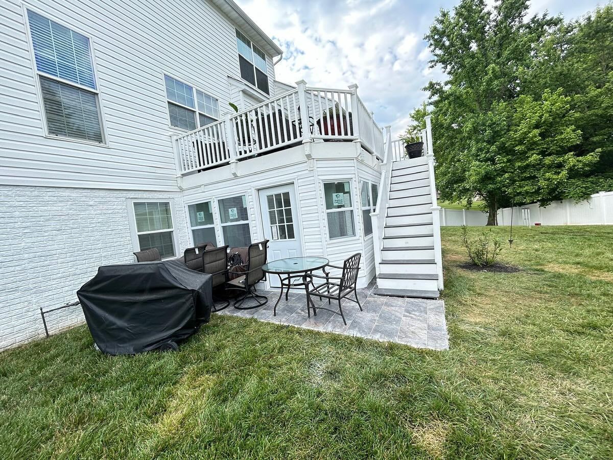 White two-story house with a deck and stairs leading to a backyard patio with a grill and patio set.