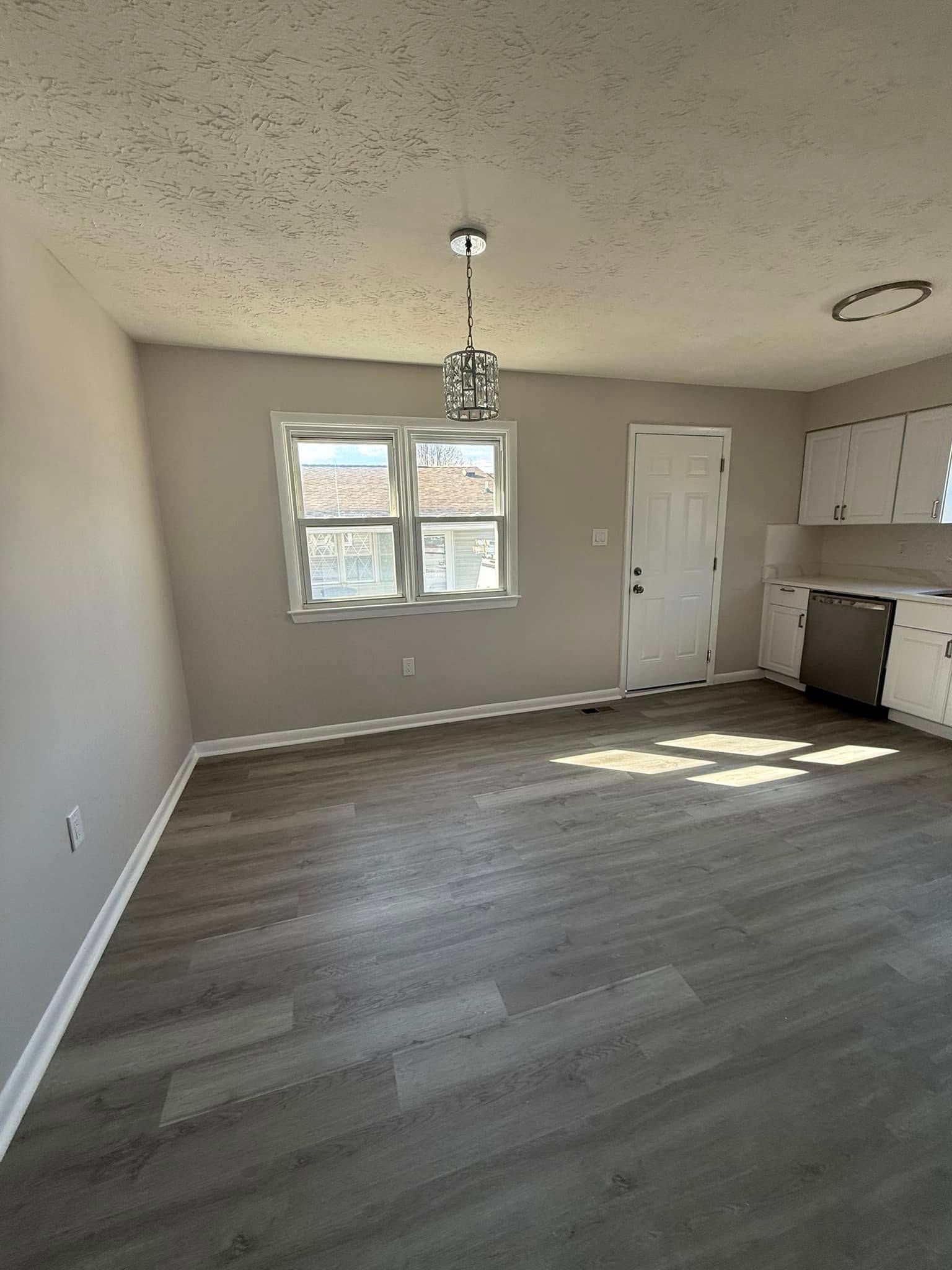 Empty room with gray flooring, tan walls, window, white cabinets, and a chandelier.