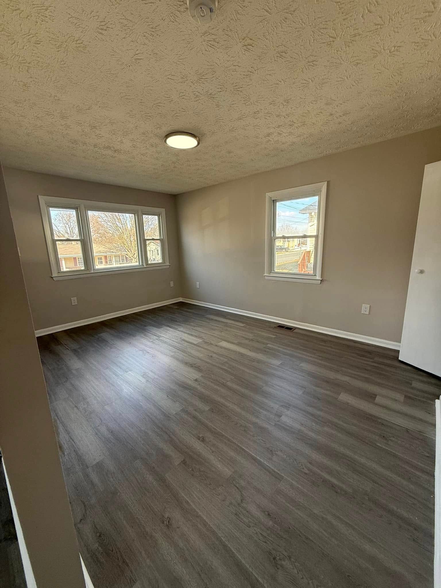 Empty room with wood-look flooring, two windows, and a ceiling light. Beige walls, white trim.