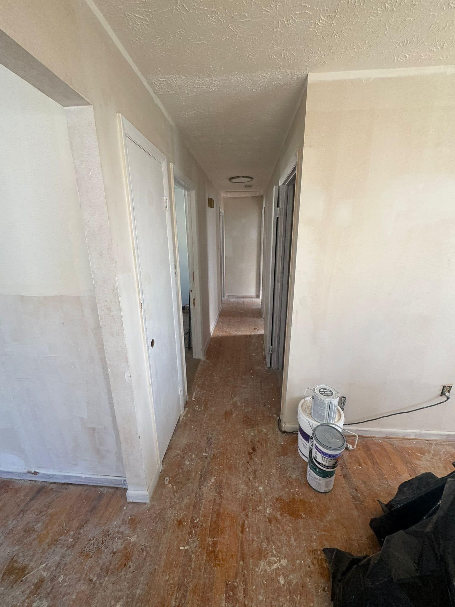 Hallway under renovation with light-colored walls and doors. Paint cans on floor, debris scattered.