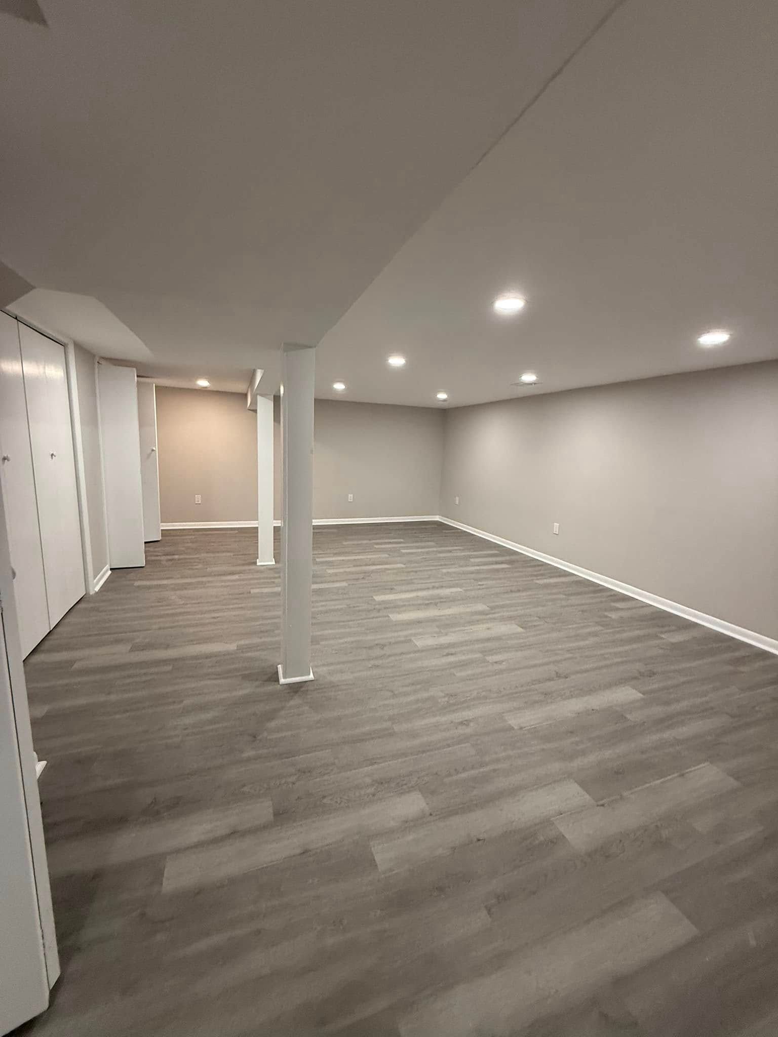Empty gray-walled basement room with recessed lighting and gray wood-look flooring. White support columns.