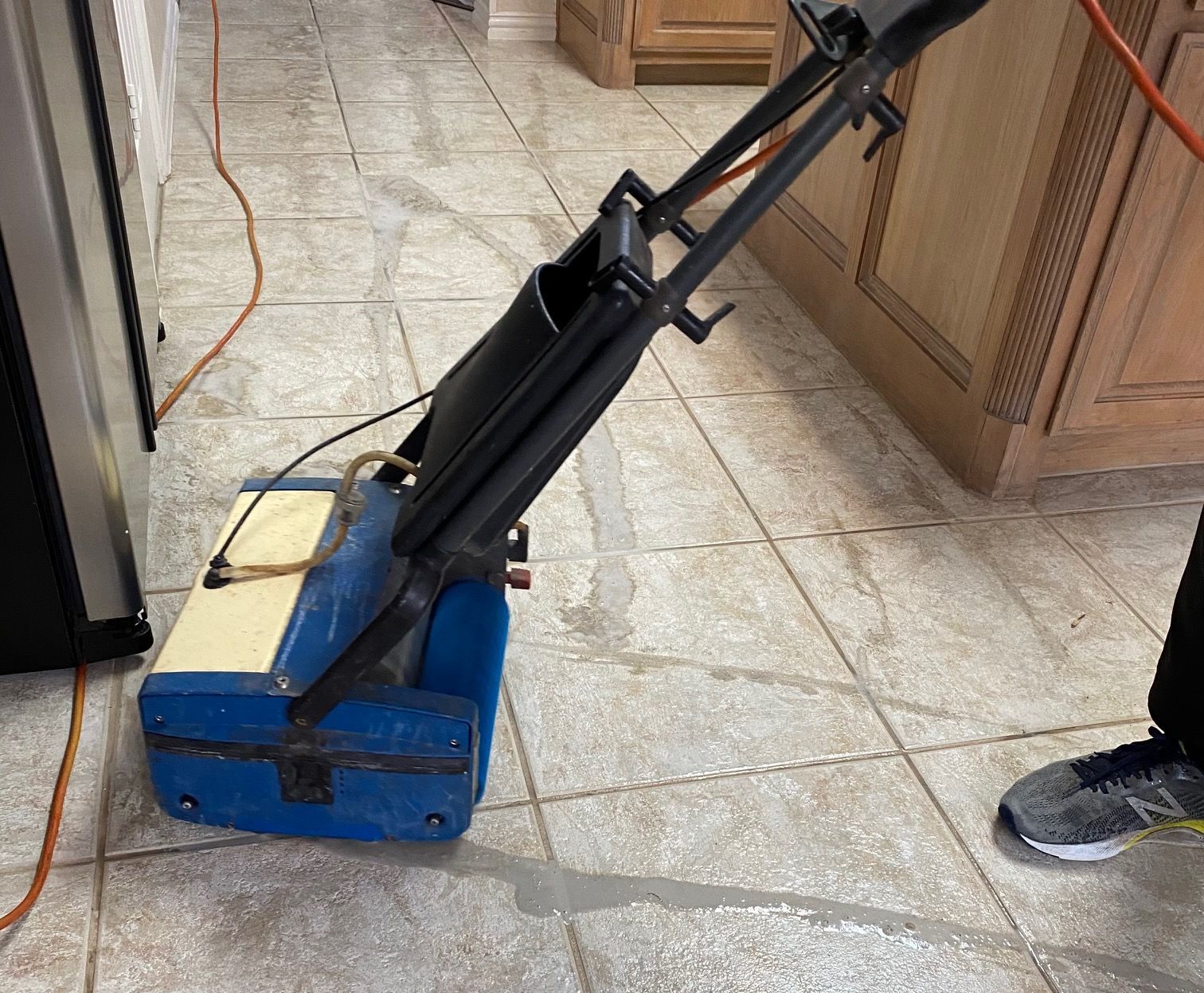 A person using a blue and beige floor cleaning machine on tile floor in a kitchen.