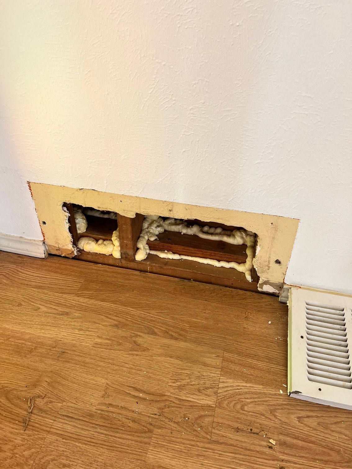 Wall vent sealed with spray foam, next to a floor vent. The floor is wood-look laminate.