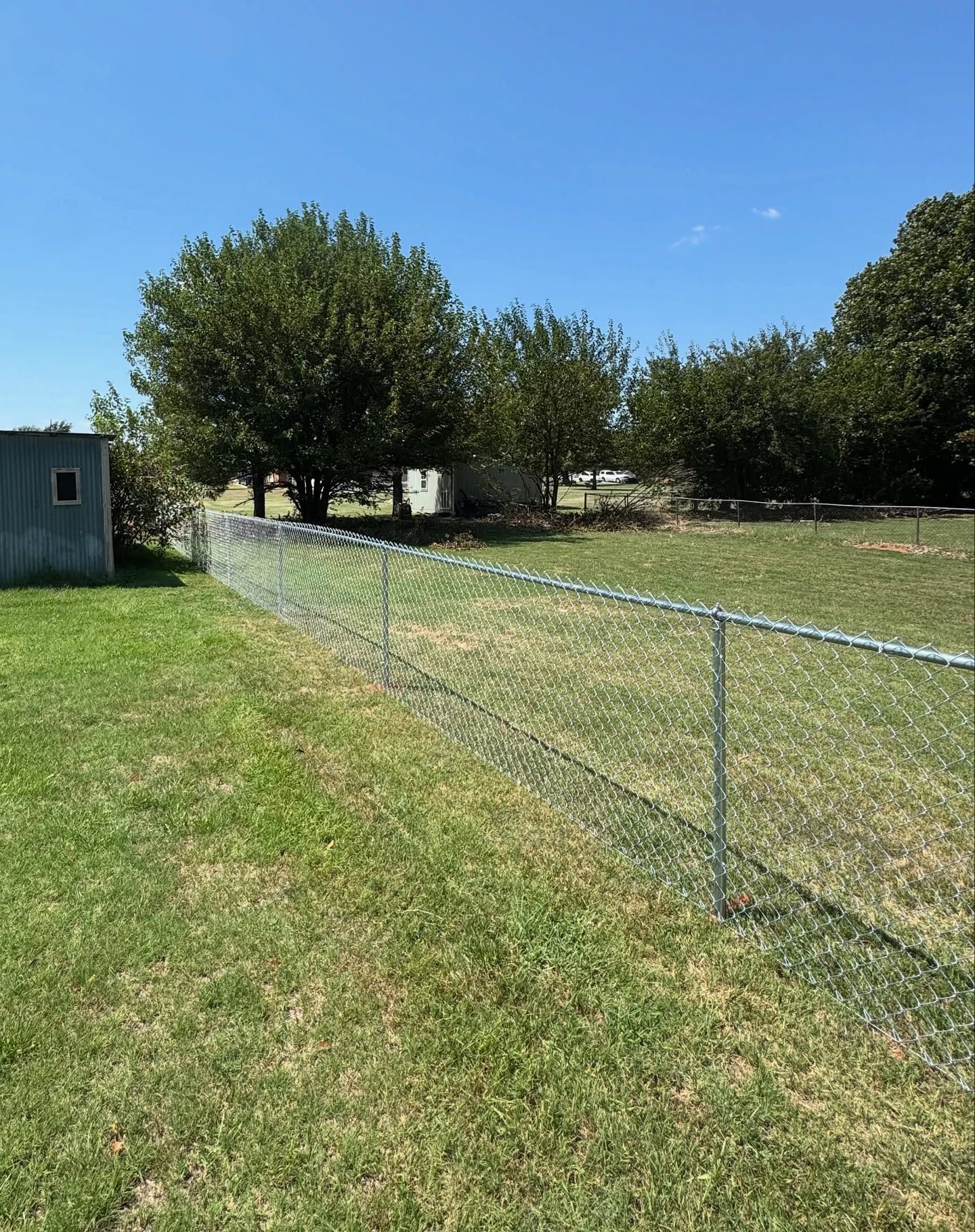 Chain-link fence in a grassy yard on a sunny day, with trees and a small building in the background. Chain-link fence in a grassy yard on a sunny day, with trees and a small building in the background.