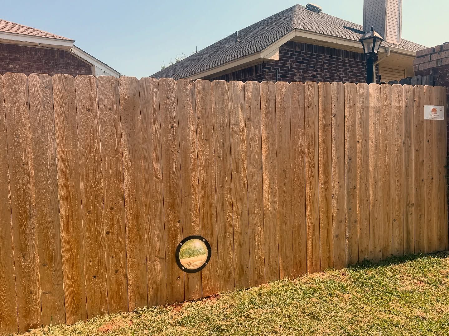 Wooden fence with a small, round window at the bottom, in front of a house and green grass. Wooden fence with a small, round window at the bottom, in front of a house and green grass.