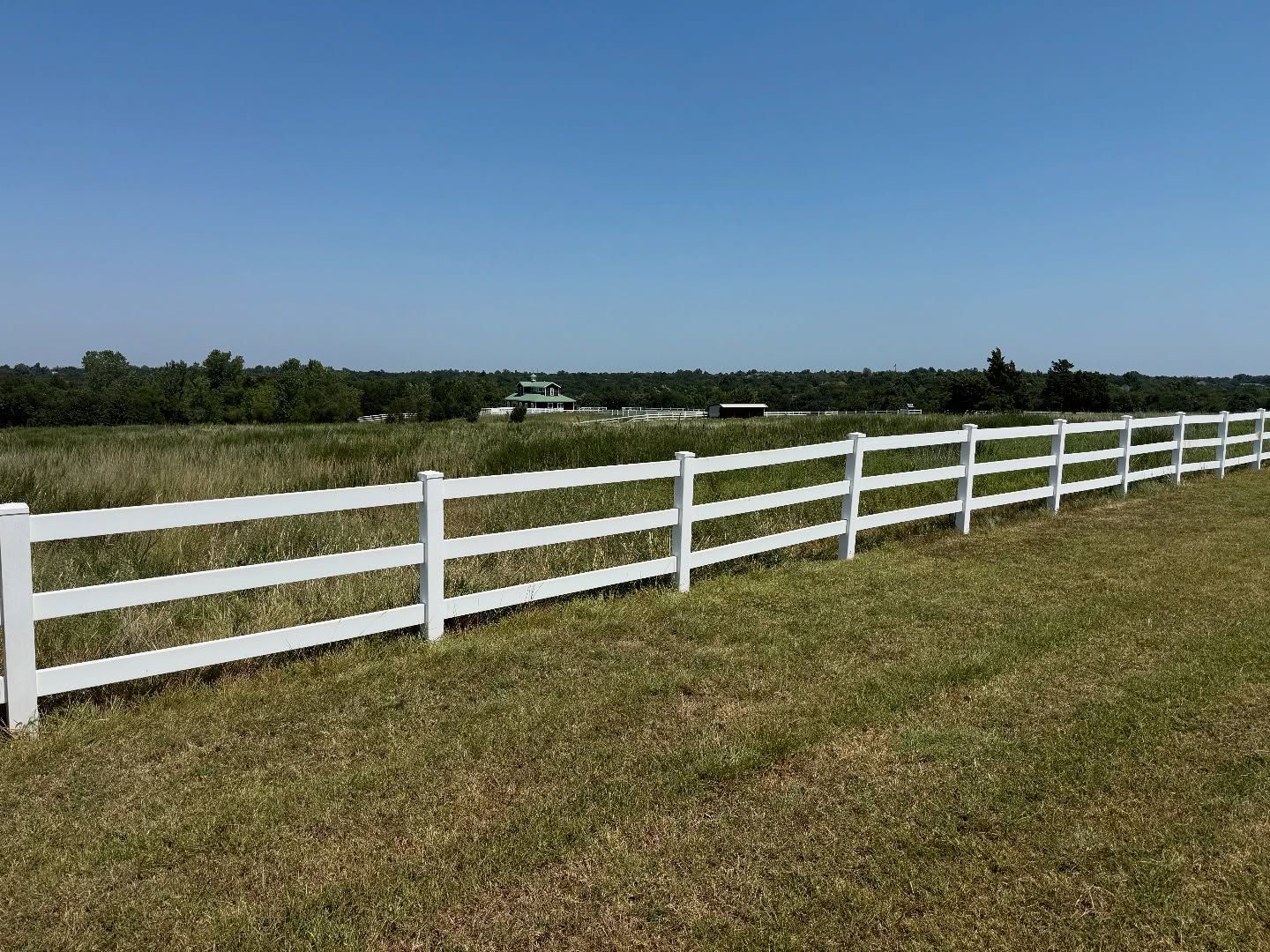 White fence in grassy field, with trees and a building in the background under a blue sky. White fence in grassy field, with trees and a building in the background under a blue sky.