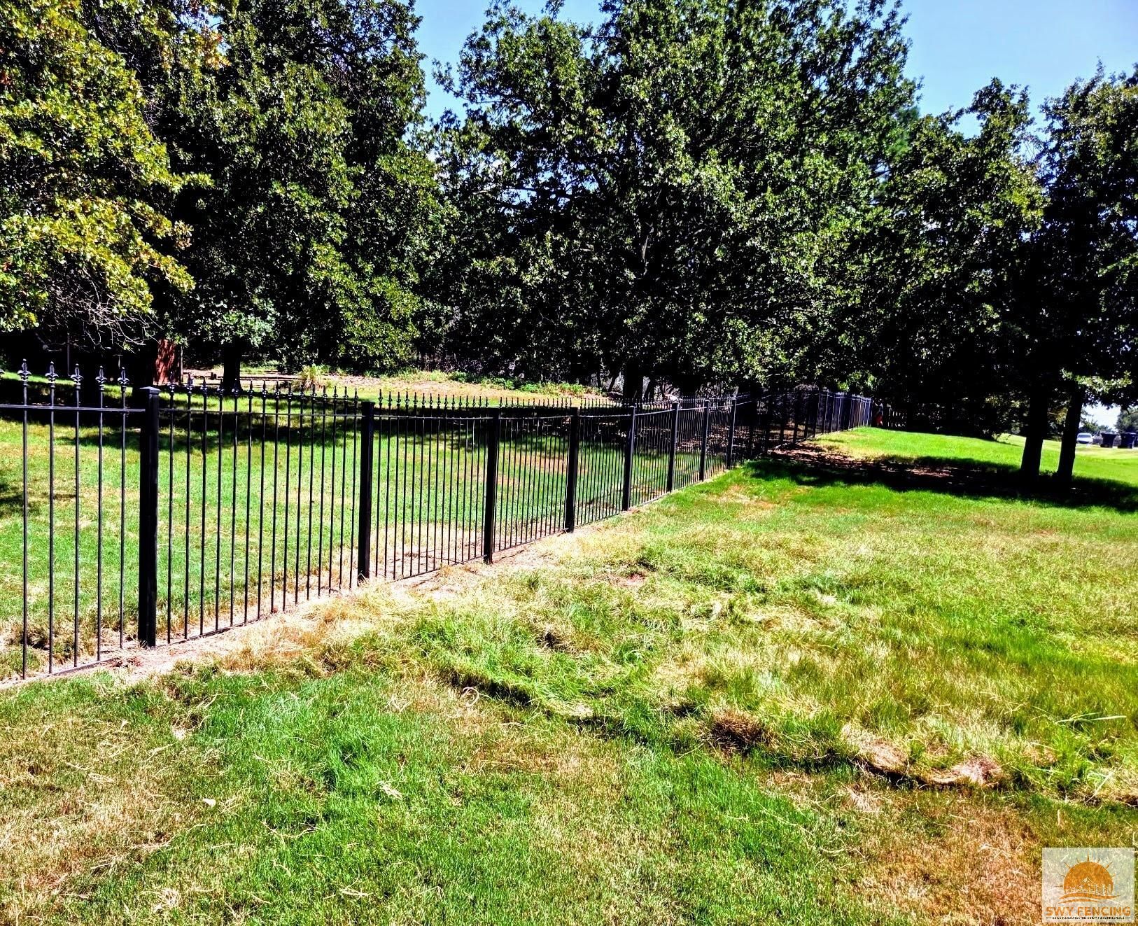Black metal fence borders a grassy area with trees on a sunny day. Black metal fence borders a grassy area with trees on a sunny day.