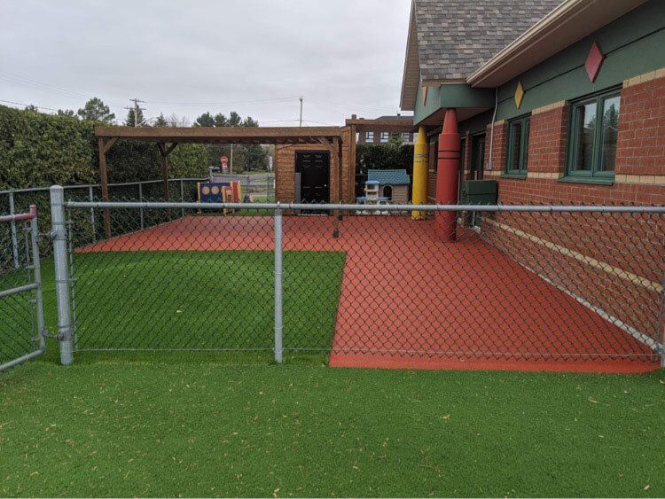 Une clôture à mailles losangées entoure une zone herbeuse devant un bâtiment en brique
