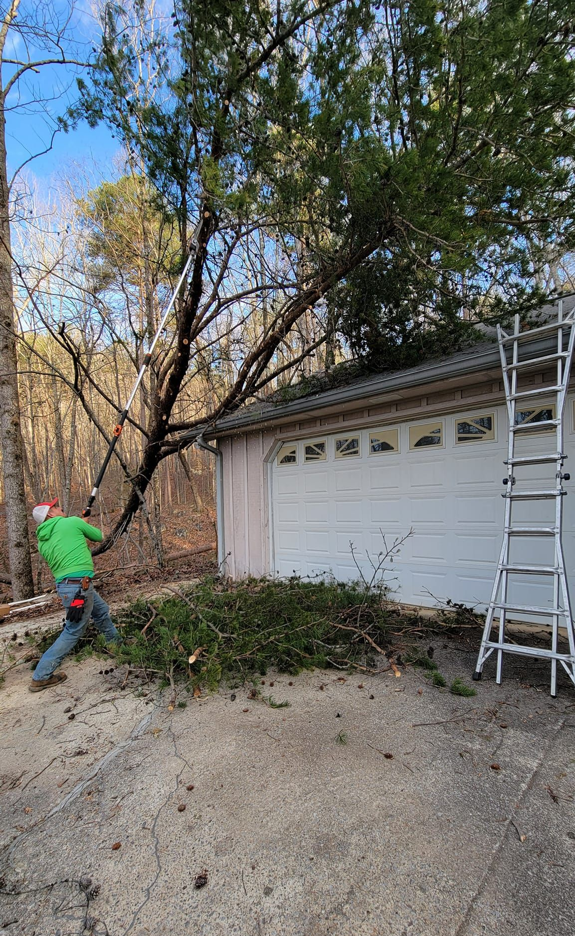 Person raking leaves beside a garage with a ladder, under a tree in a yard.