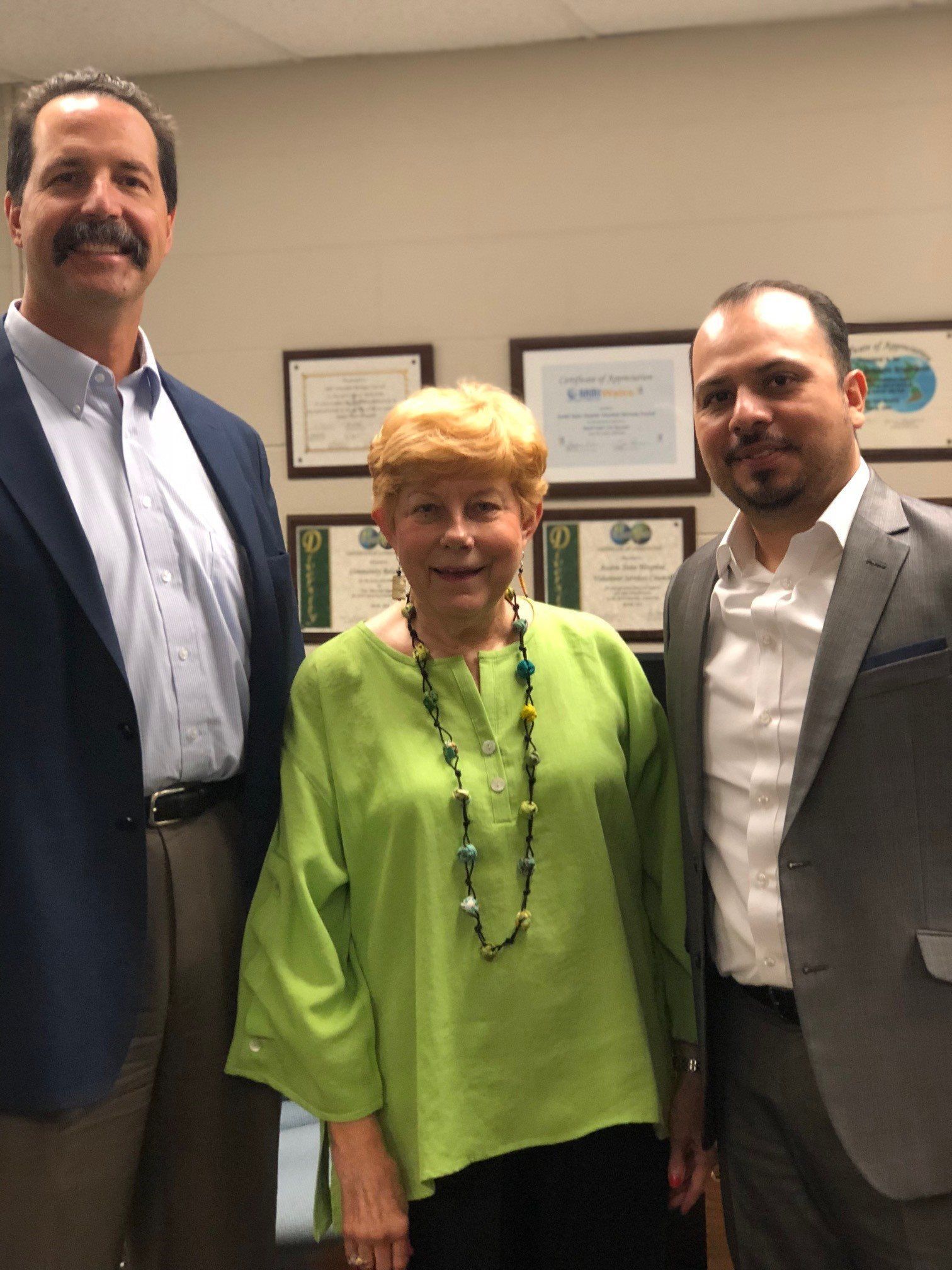 Three people standing in an office. A woman in lime green top is centered between two men in suits.