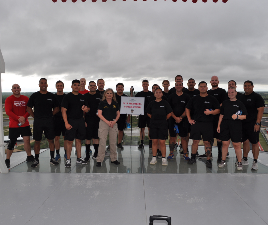 Group of people standing on a glass platform. Some in black shirts, one in tan. Sign in center. Cloudy sky.