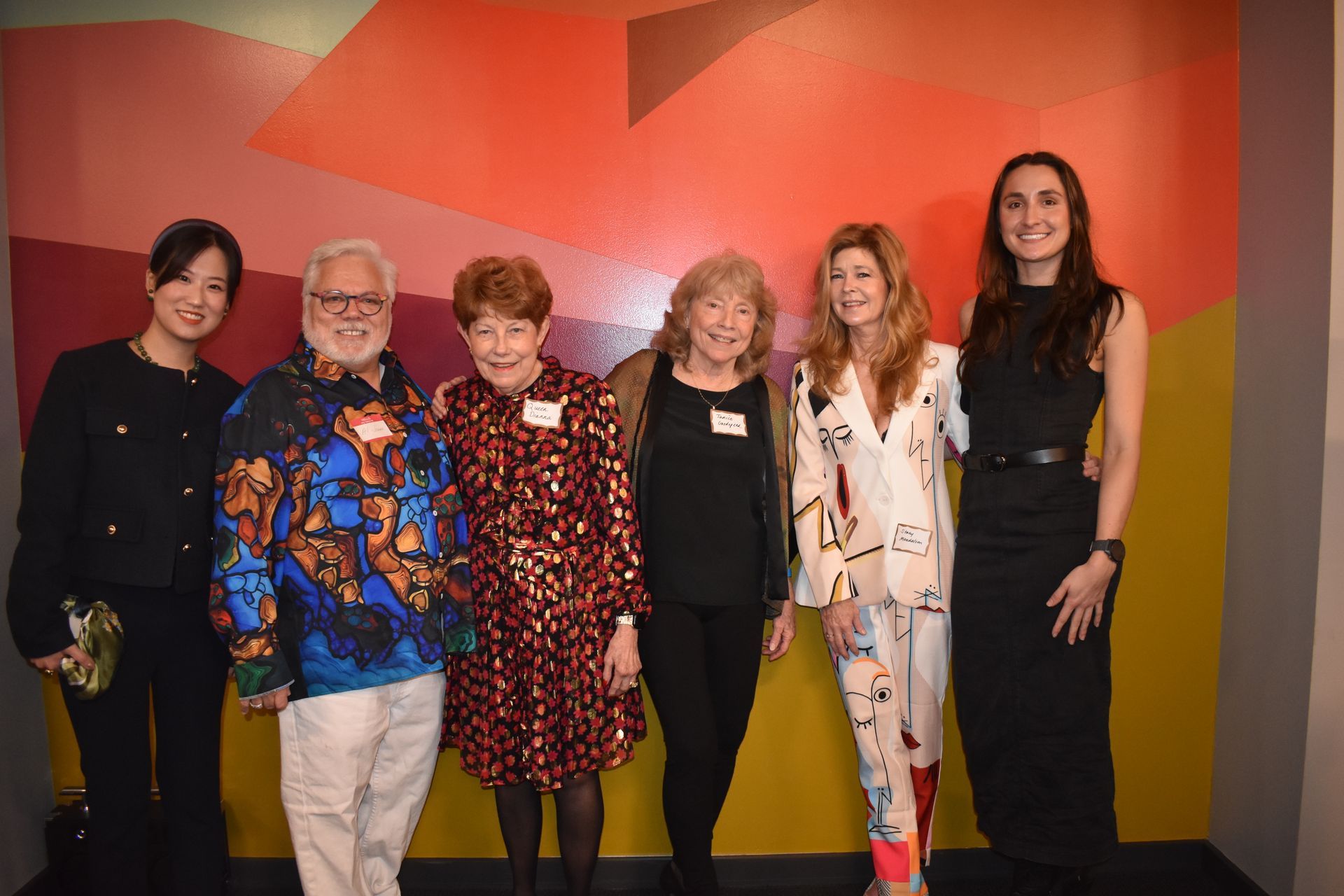 Six people pose in front of a colorful wall; one man wears a patterned shirt, others wear neutral colors.