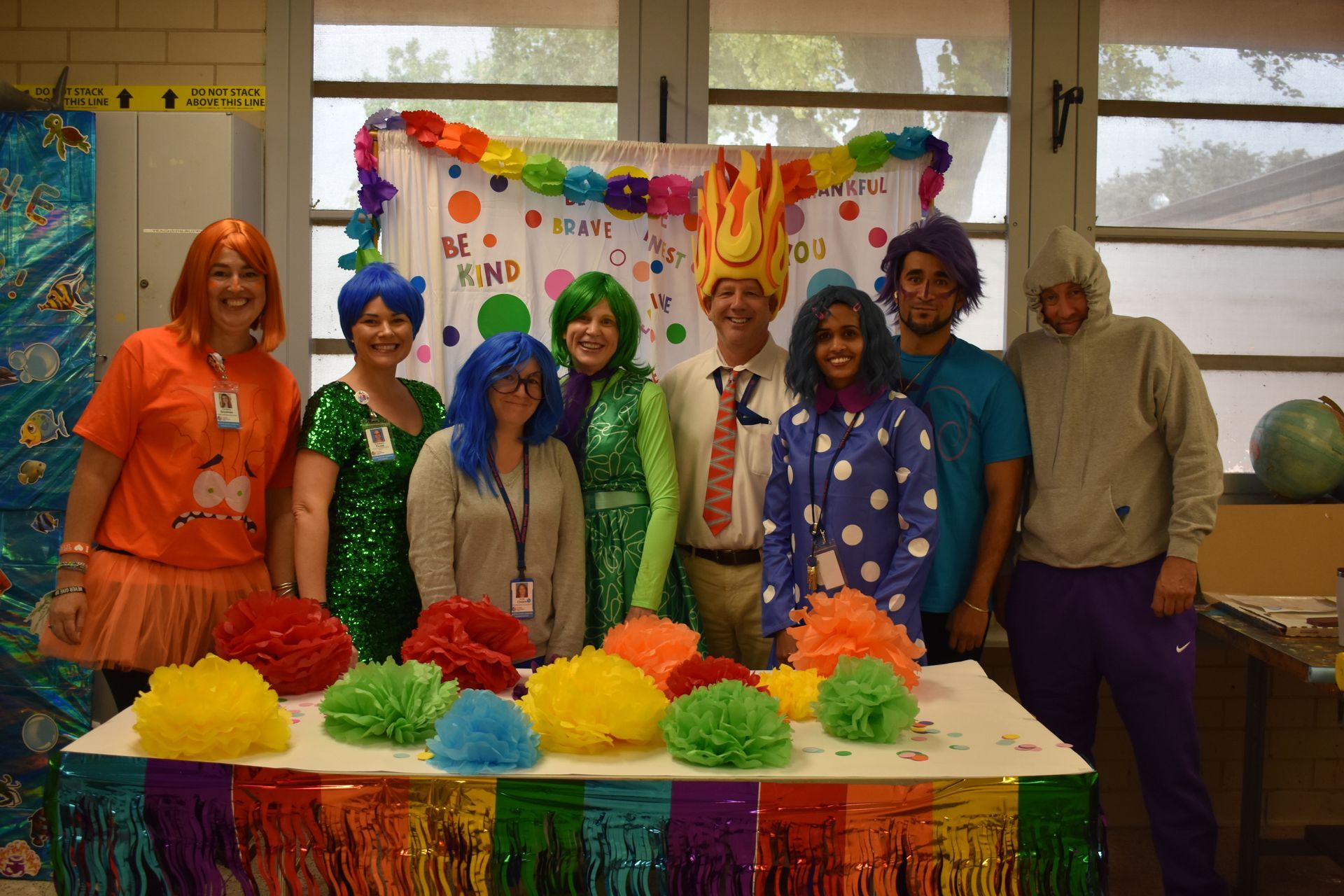 Group of people posing behind colorful pom-poms and a rainbow banner at a festive indoor event.
