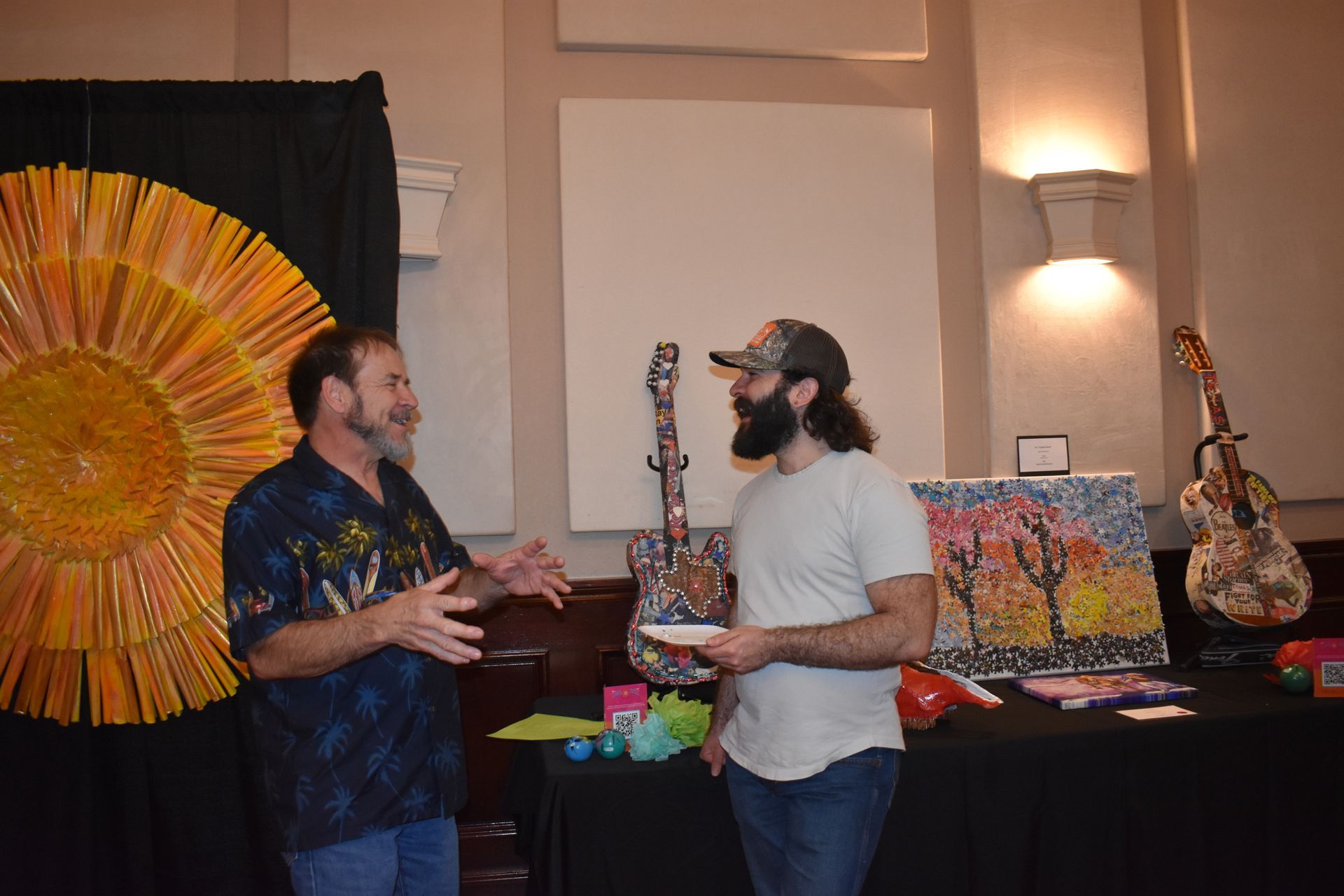 Two men talking near art display; one gestures, the other holds a plate. Guitars and paintings on the table, sun-shaped art on the wall.
