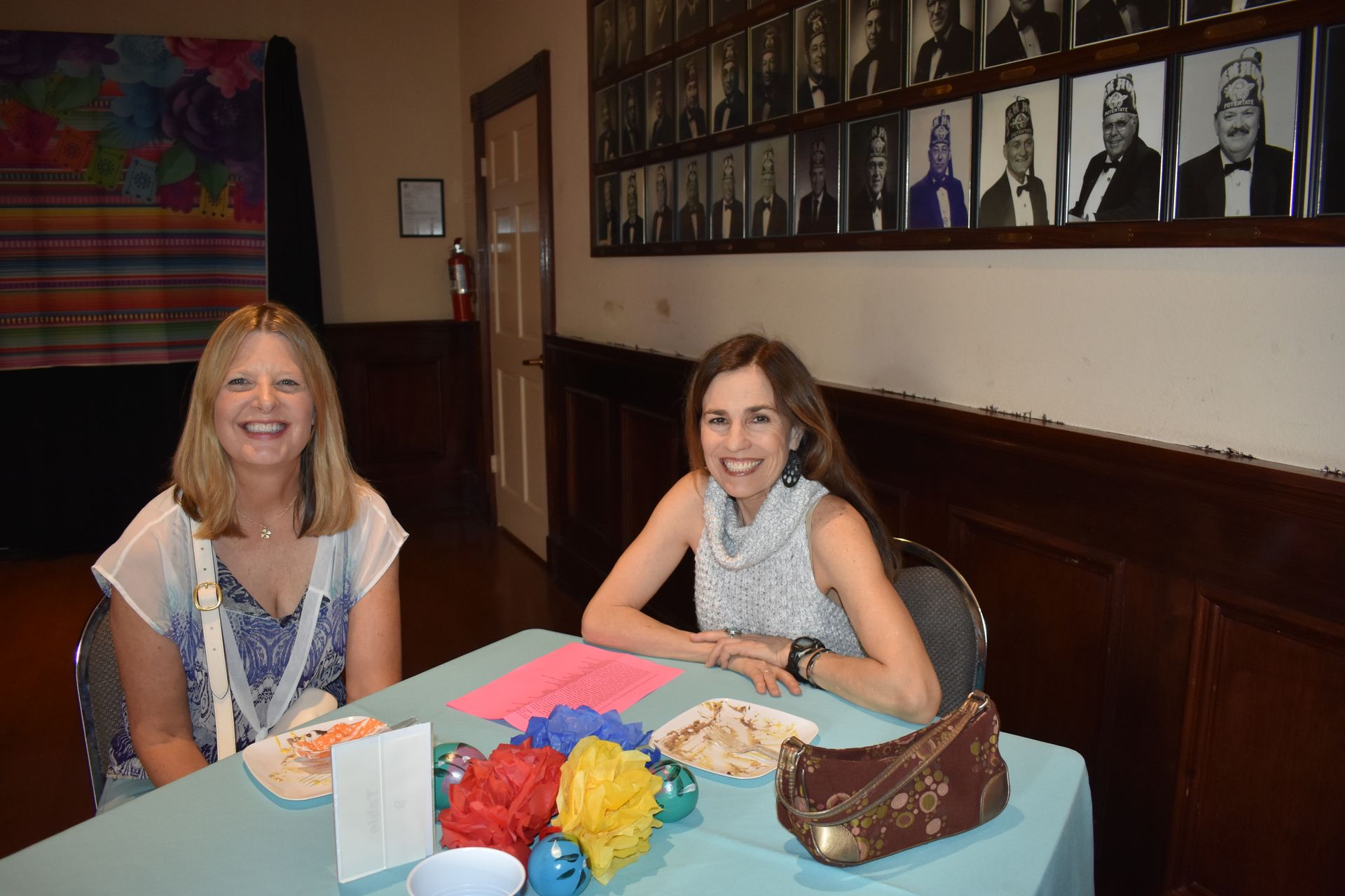 Two women smile at a table with snacks and decorations; photos hang on the wall.
