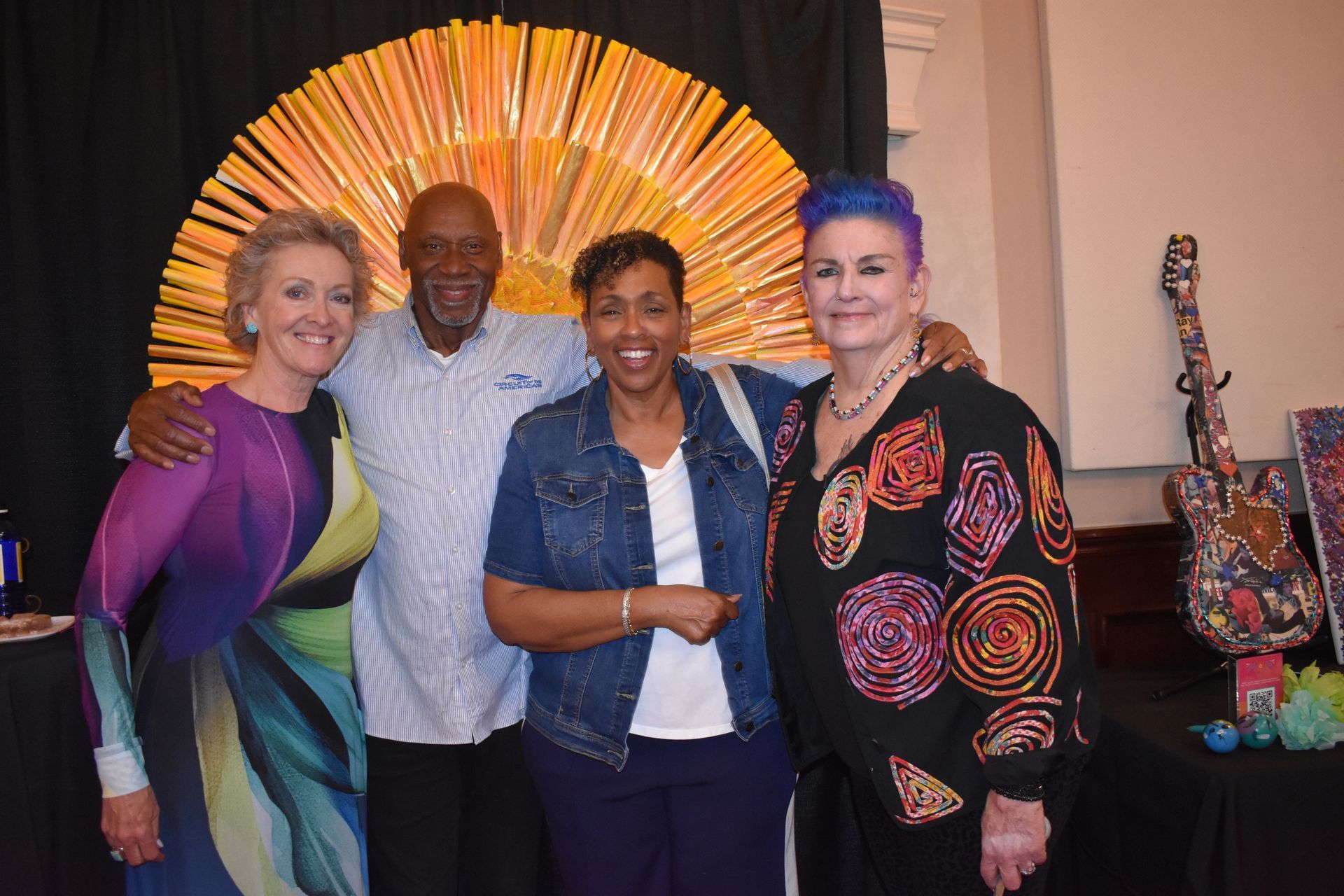 Four people pose in front of a sun-shaped art piece. One woman wears purple and the other a patterned top; two men are also in the photo.