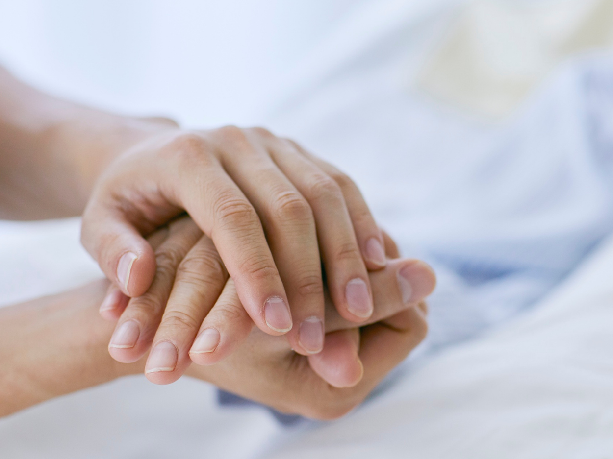 Two hands clasped together, possibly offering comfort, in a hospital bed setting.